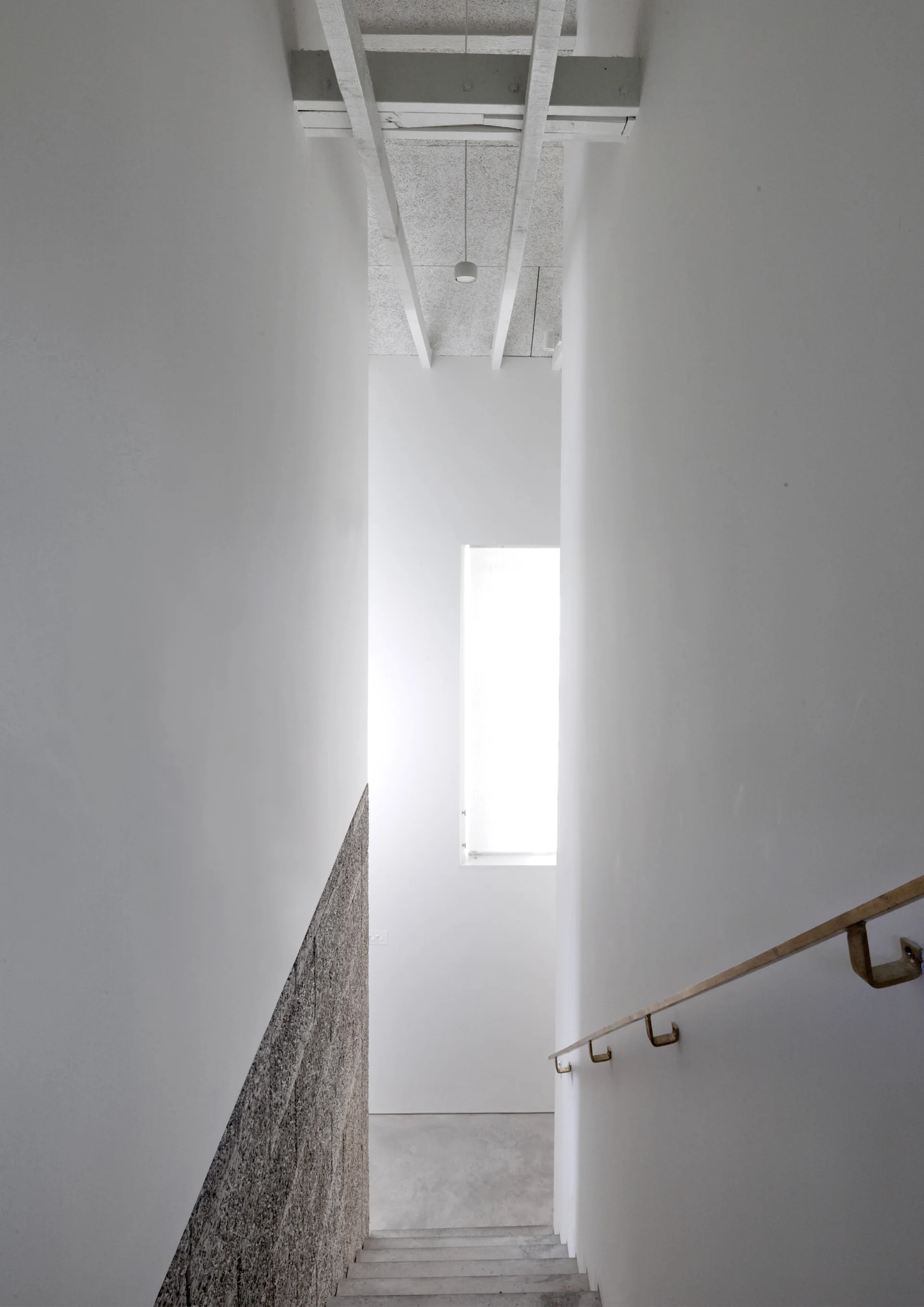 A vertical view of a modern minimalist stairwell featuring smooth white walls, concrete steps, a bronze handrail, and a textured lower wall section.