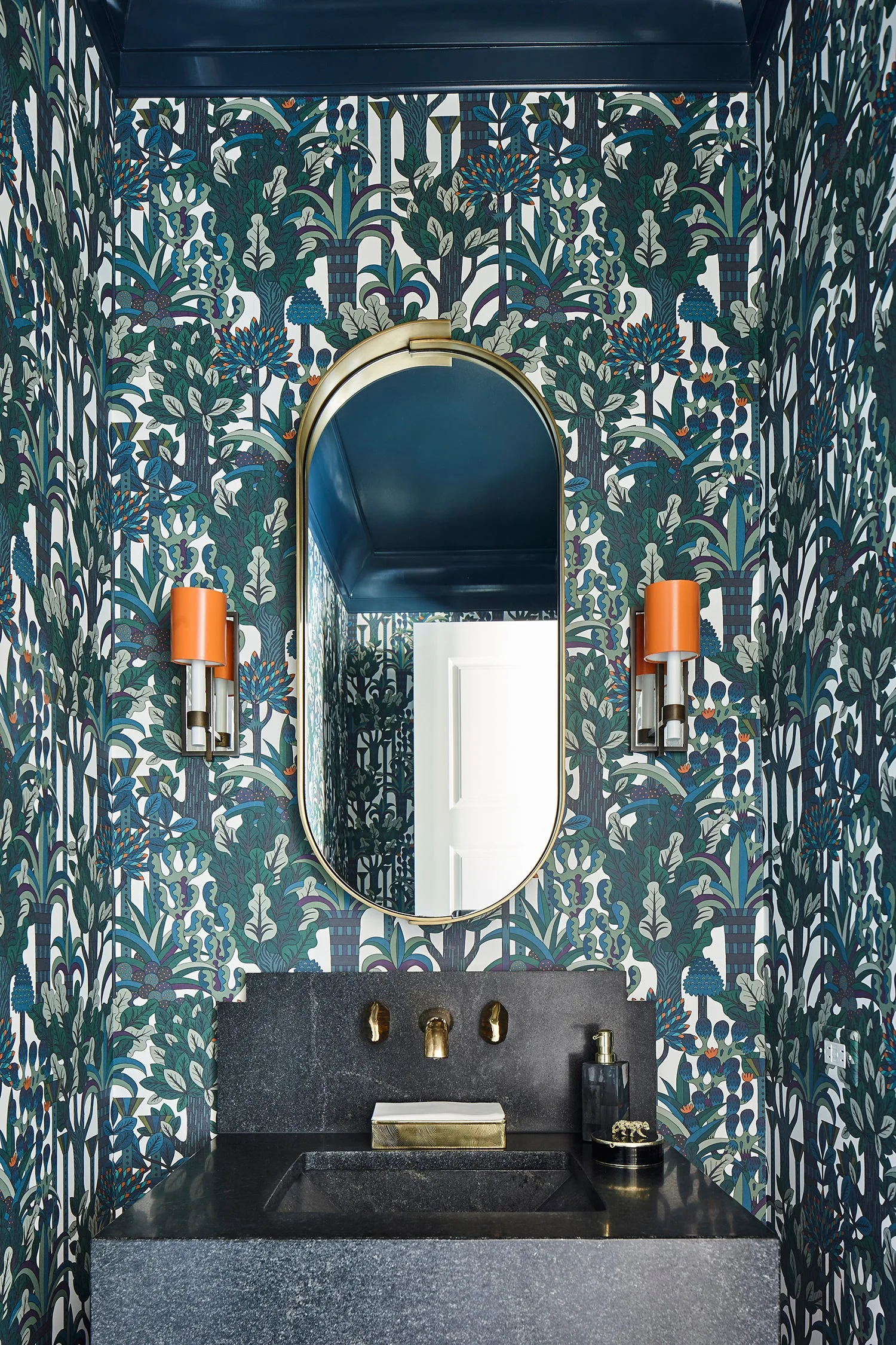 A bold powder room featuring green botanical wallpaper, an oval brass mirror, orange sconces, and a black stone vanity with a brass faucet.