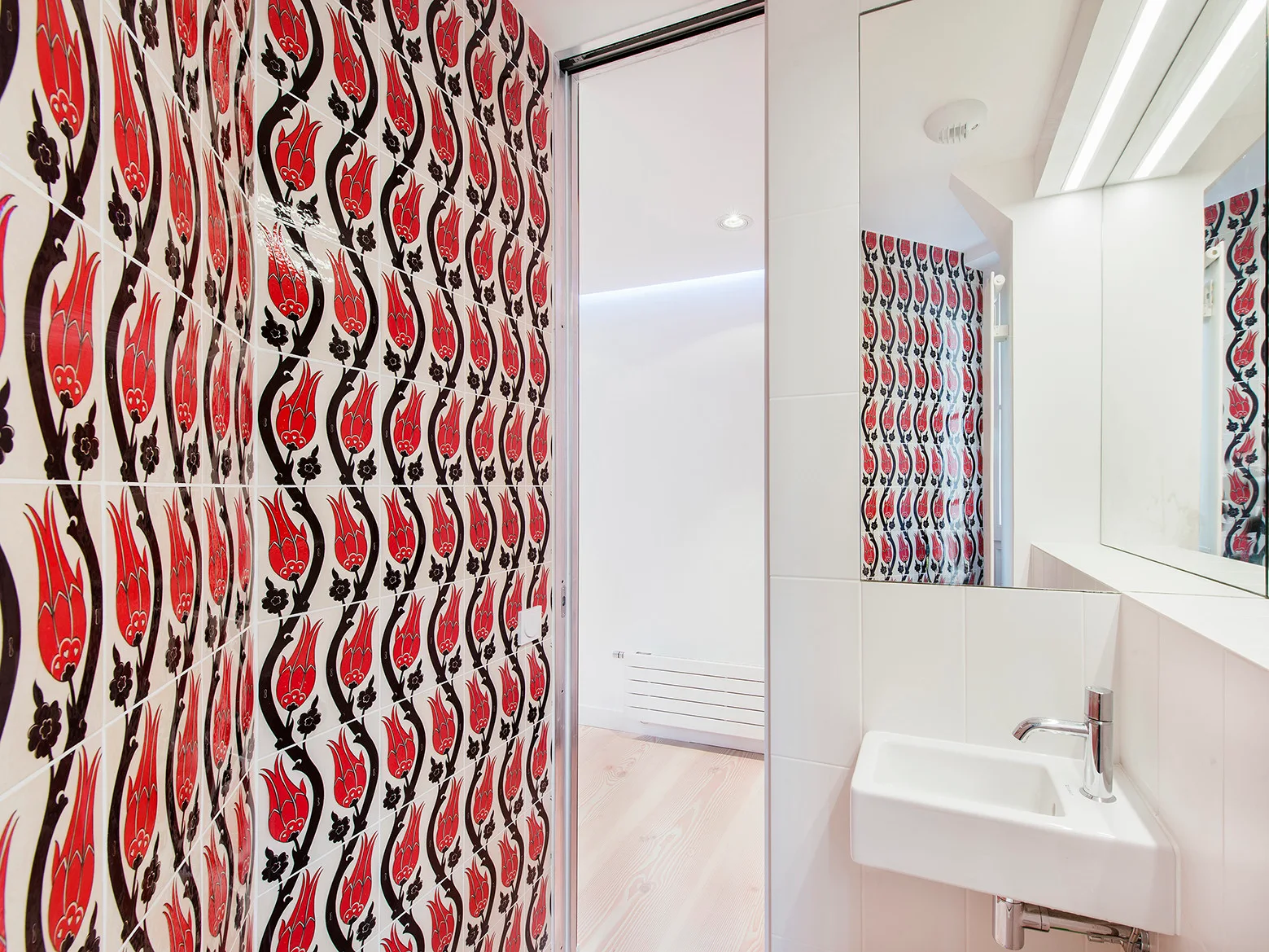 A bathroom featuring a wall tiled in a red and black tulip pattern next to a white floating sink and a large wall mirror.