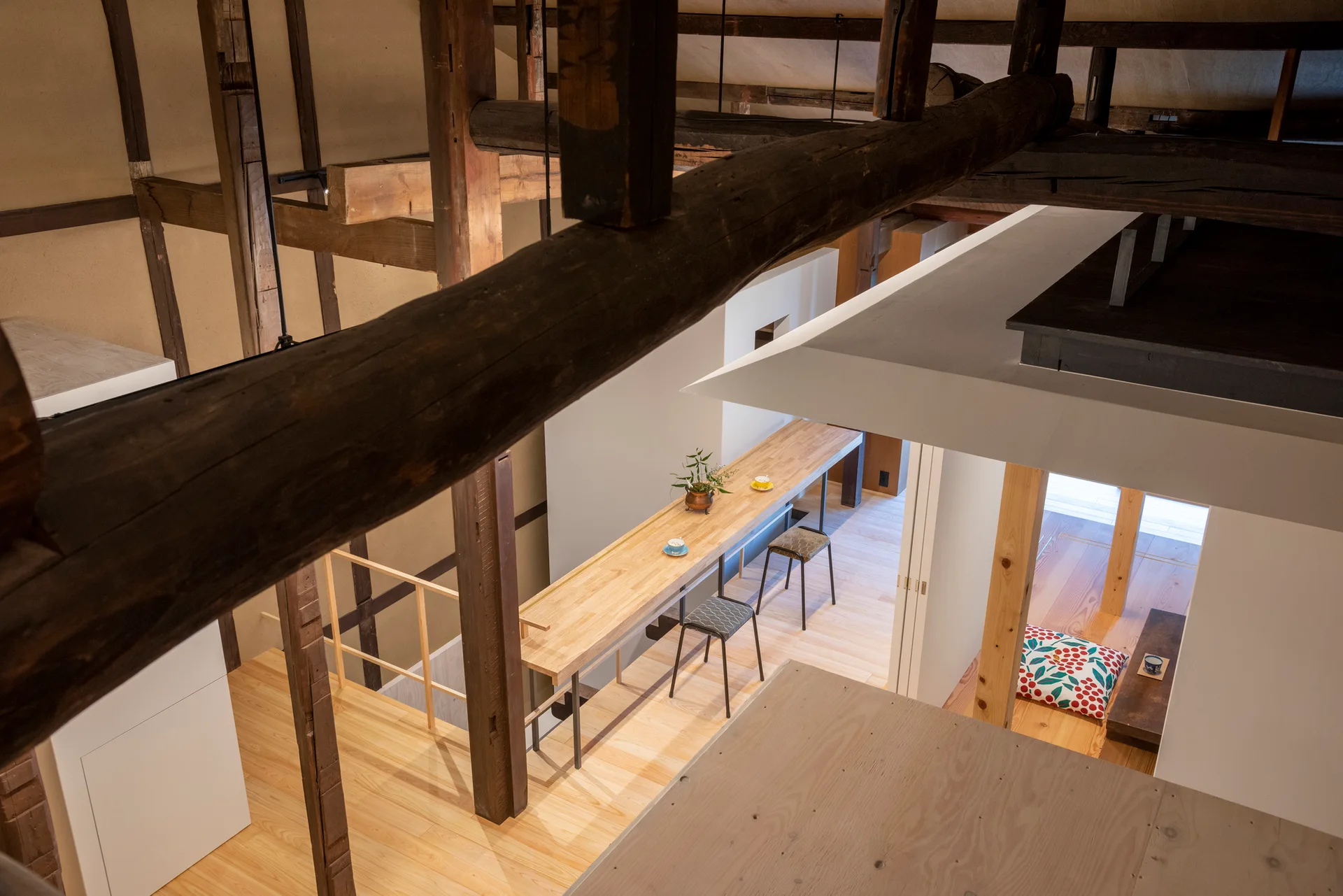 High-angle view of a loft interior featuring dark exposed timber ceiling beams, a long light wood bar table with two stools, and white partition walls.