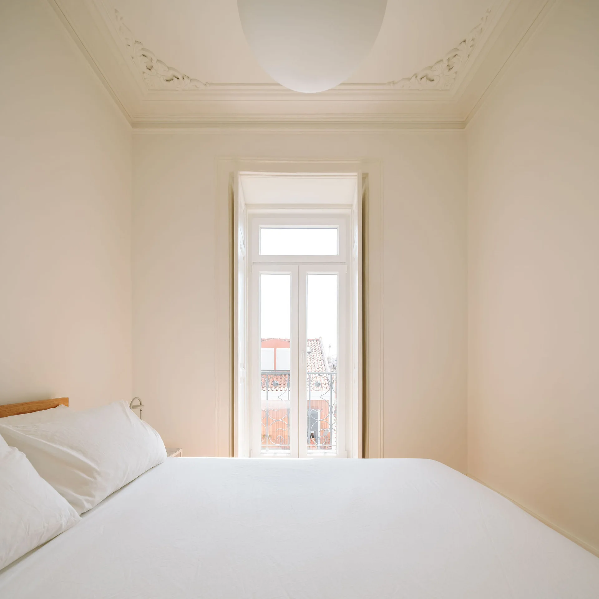 An off-white minimalist bedroom featuring a bed with white linens beneath an ornate plaster crown moulding and a tall central window.