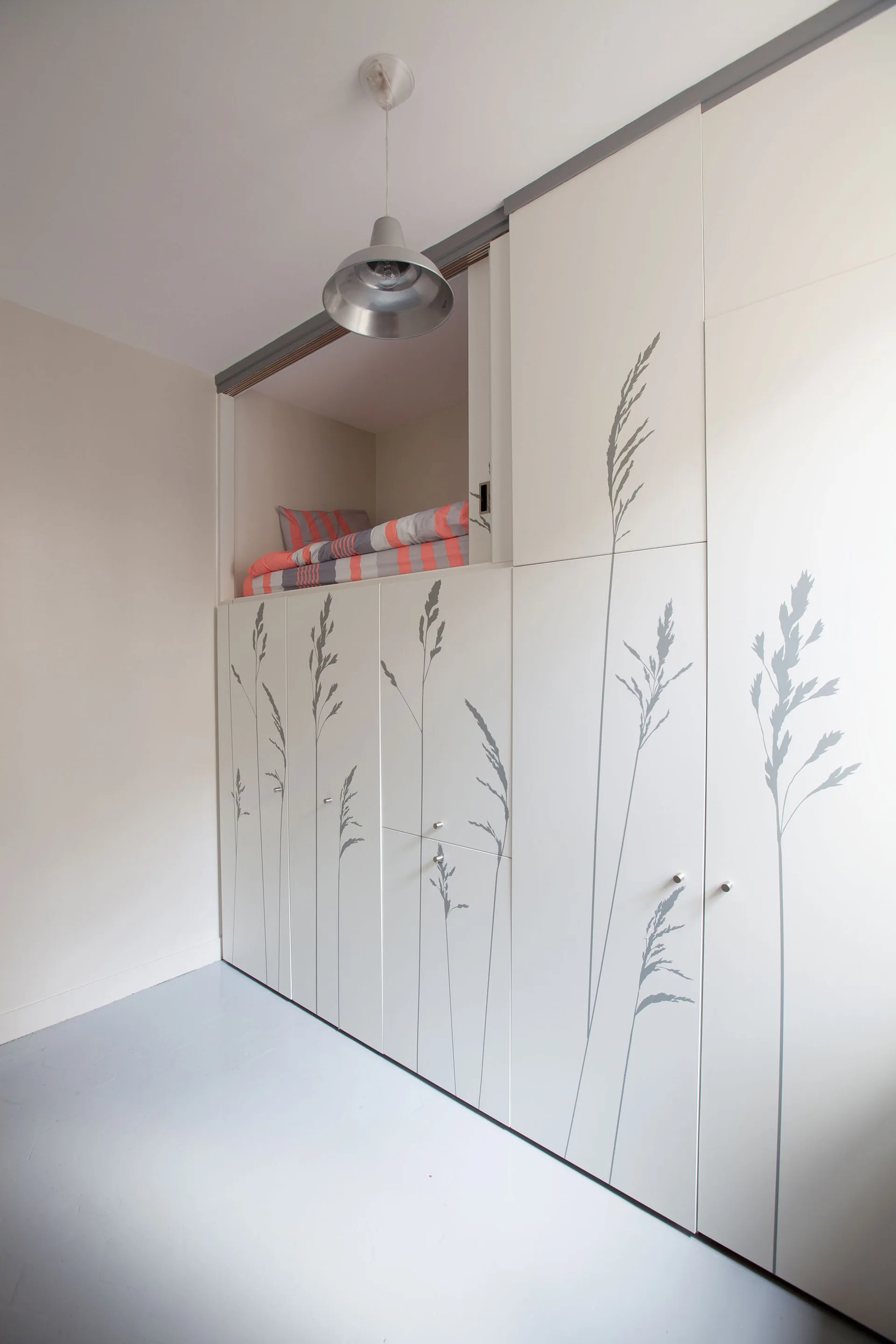 A compact bedroom features a custom white wardrobe unit with botanical grass decals and an integrated upper sleeping loft with striped bedding.