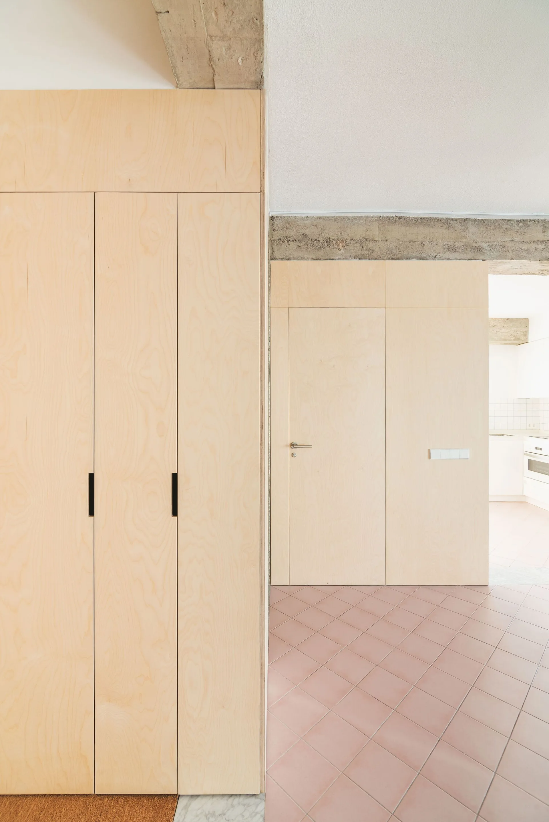 Light wood plywood cabinetry and a flush door line a hallway featuring muted pink diagonal floor tiles and an exposed concrete beam.