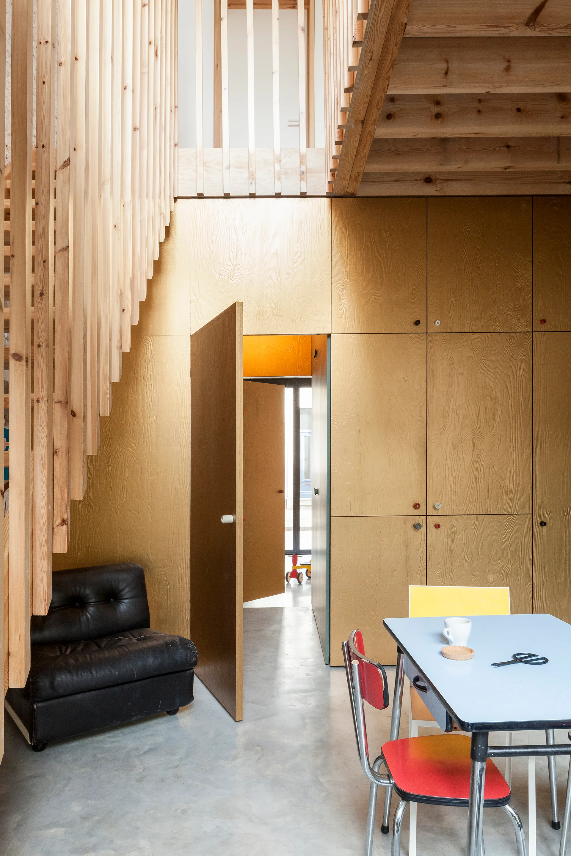 A modern interior space features light plywood wall cabinetry, a slatted wooden staircase, a black tufted chair, and a light blue dining table.