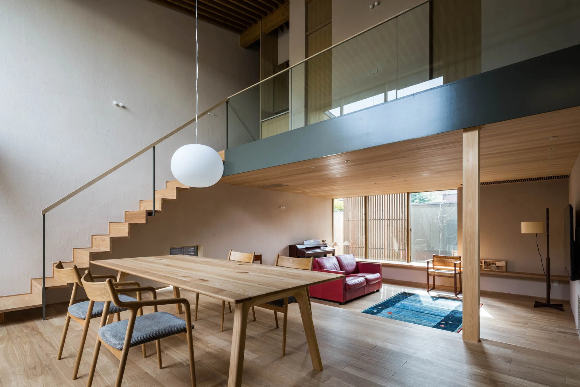 A minimalist double-height dining room features a light wood table, a floating wooden staircase, and a mezzanine with a glass railing.