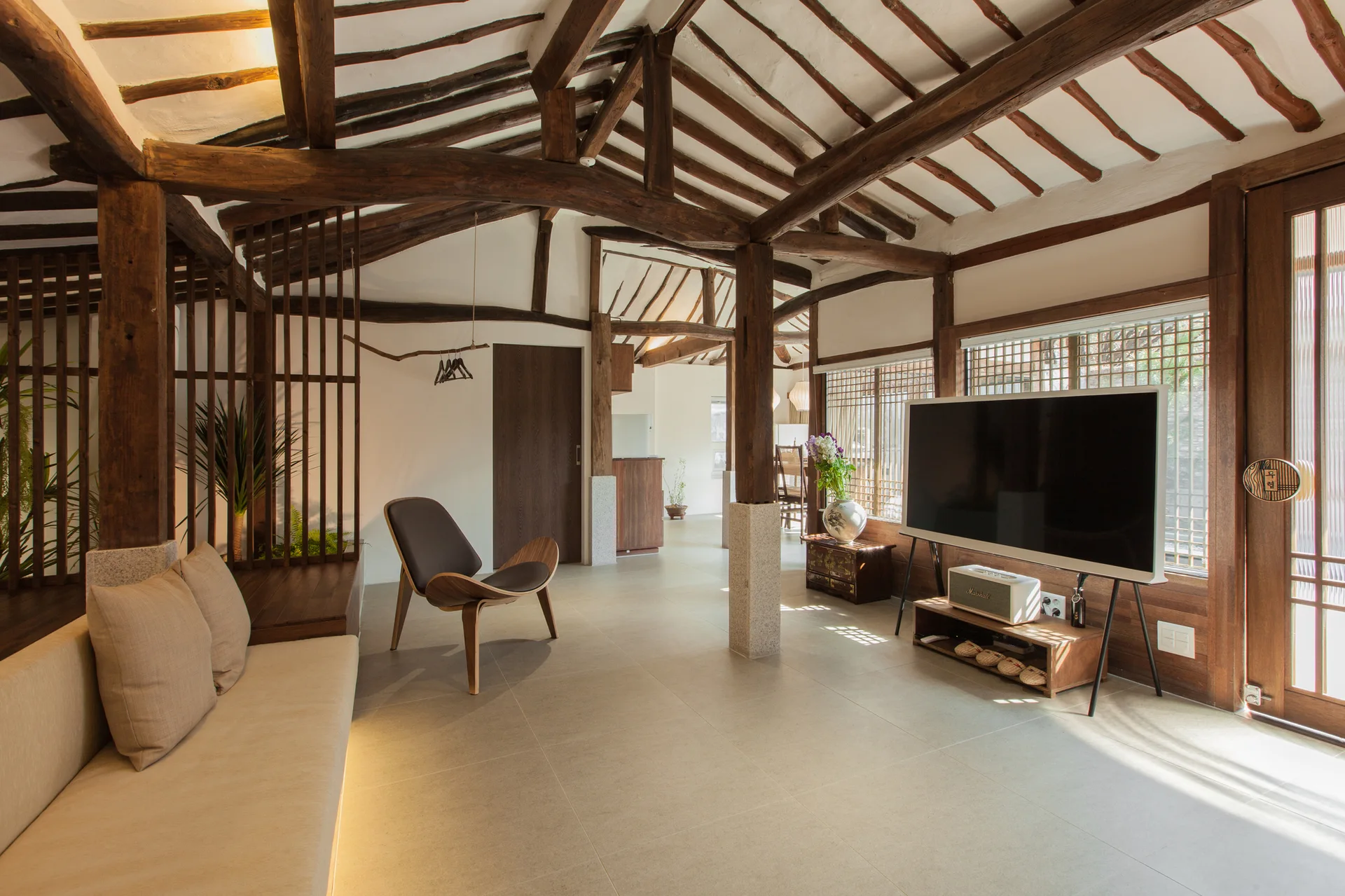 A minimalist living room with exposed dark wood rafters, a light beige sofa, a modern armchair, and a flat-screen television on a tripod stand.