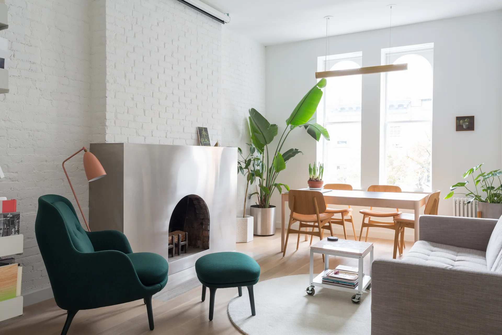 A modern living room featuring a brushed metal fireplace surround against a painted white brick wall, a green armchair, and a wood dining set.