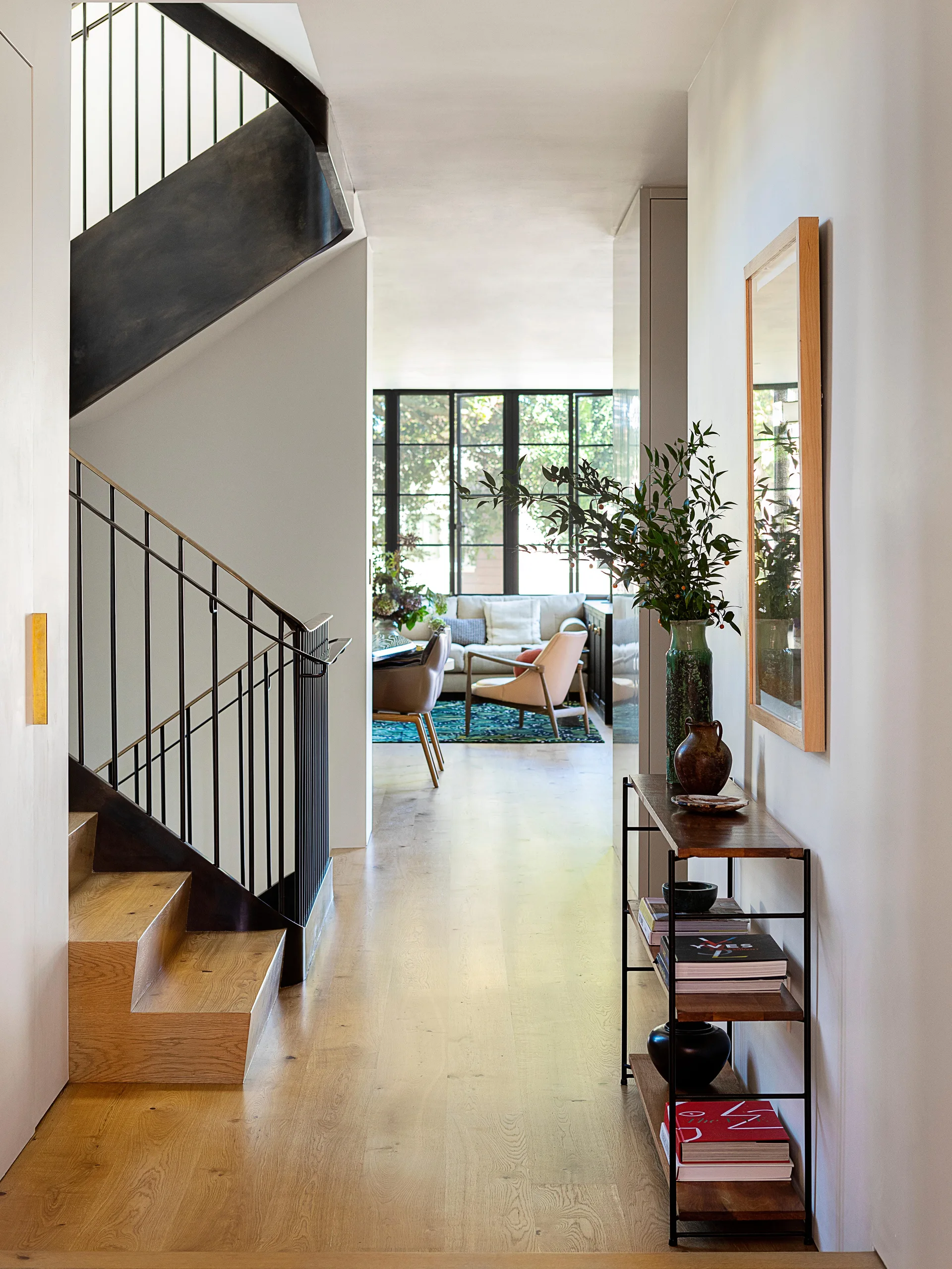 A bright hallway features light wood floors, a staircase with a black metal railing, and a slender console table beneath a wood-framed mirror.