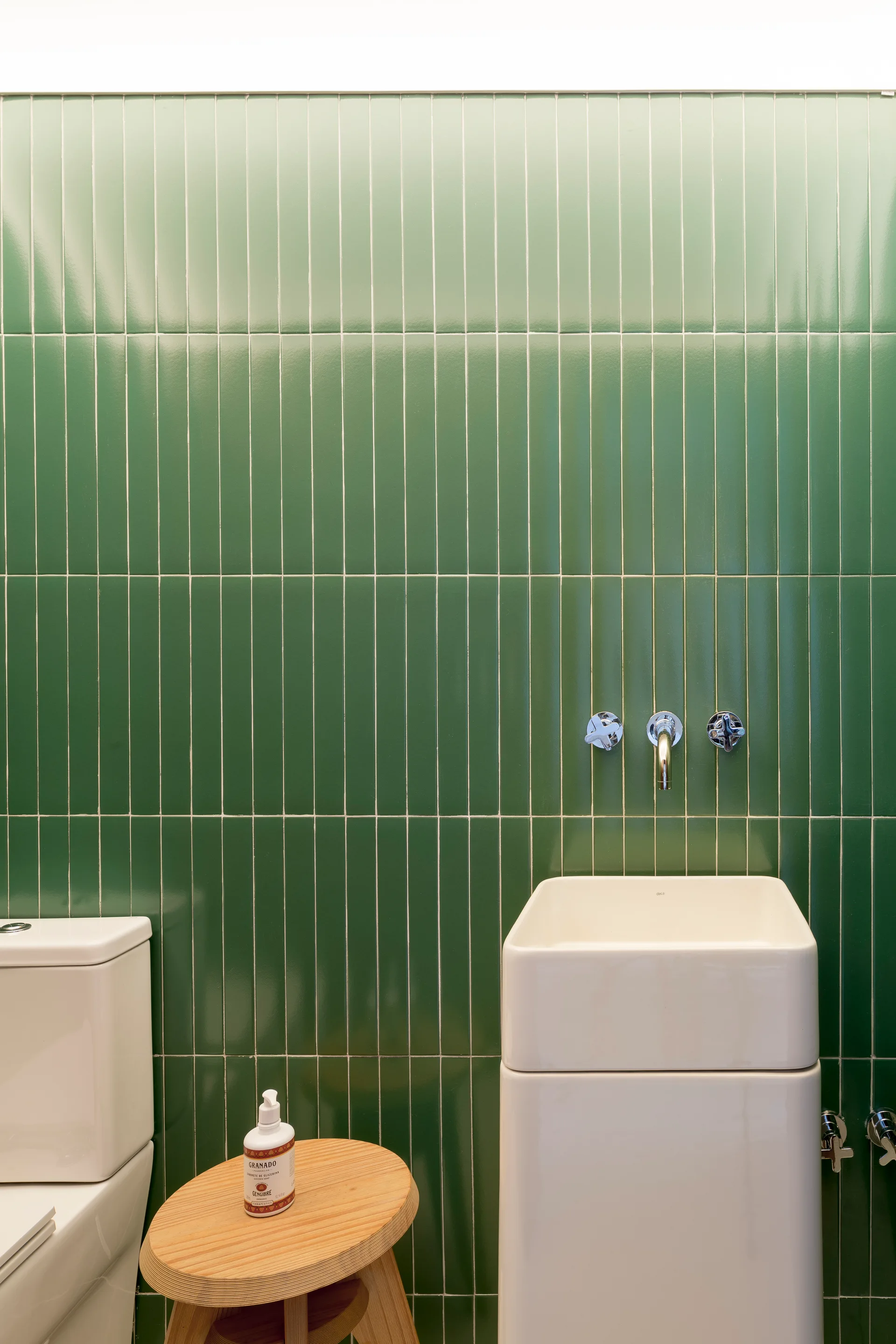 A minimalist bathroom features glossy green vertical stacked wall tiles, a white pedestal sink with a wall-mounted faucet, and a small round wood stool.