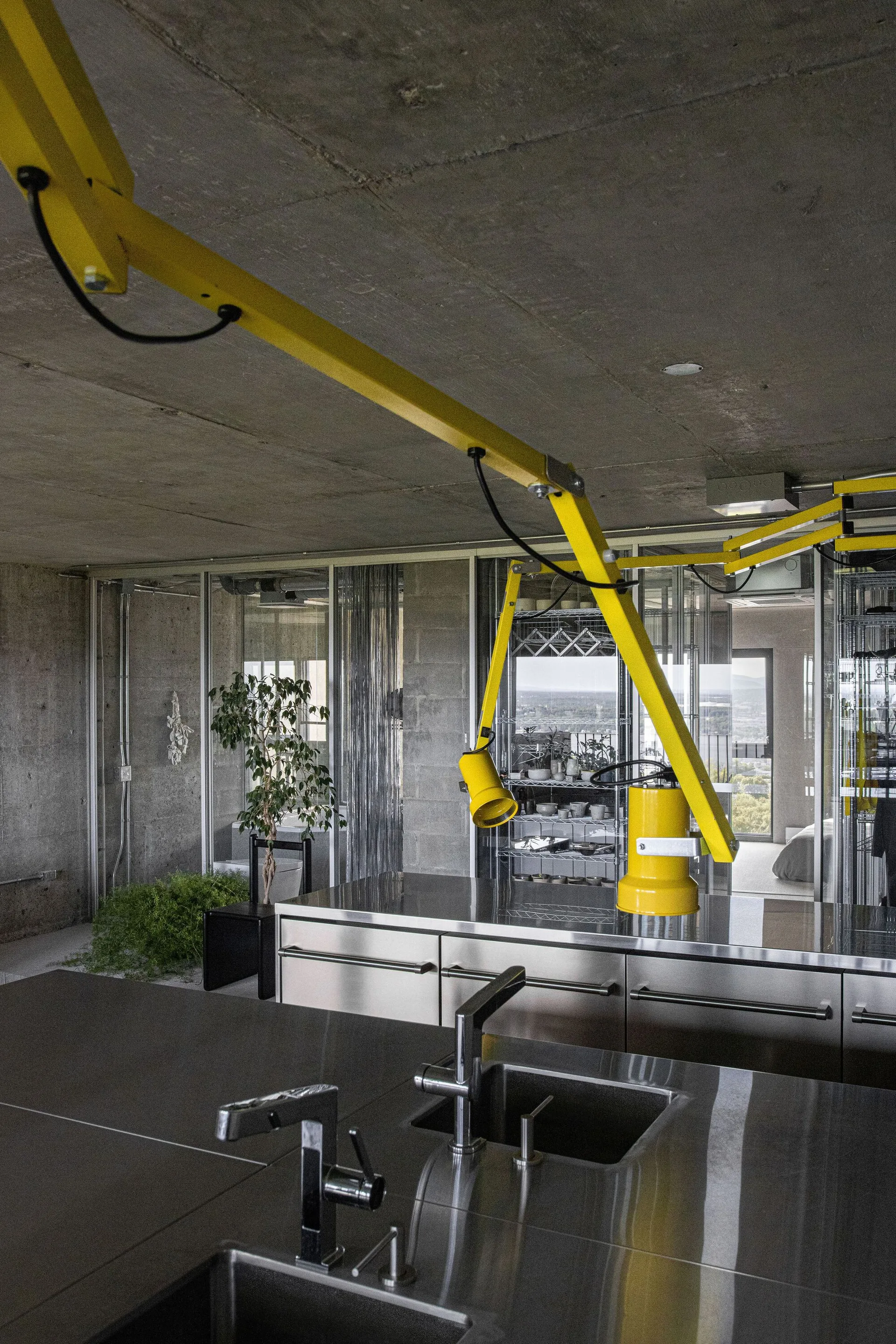 An industrial kitchen with a stainless steel island sink and raw concrete walls is lit by an overhead yellow articulating light fixture.