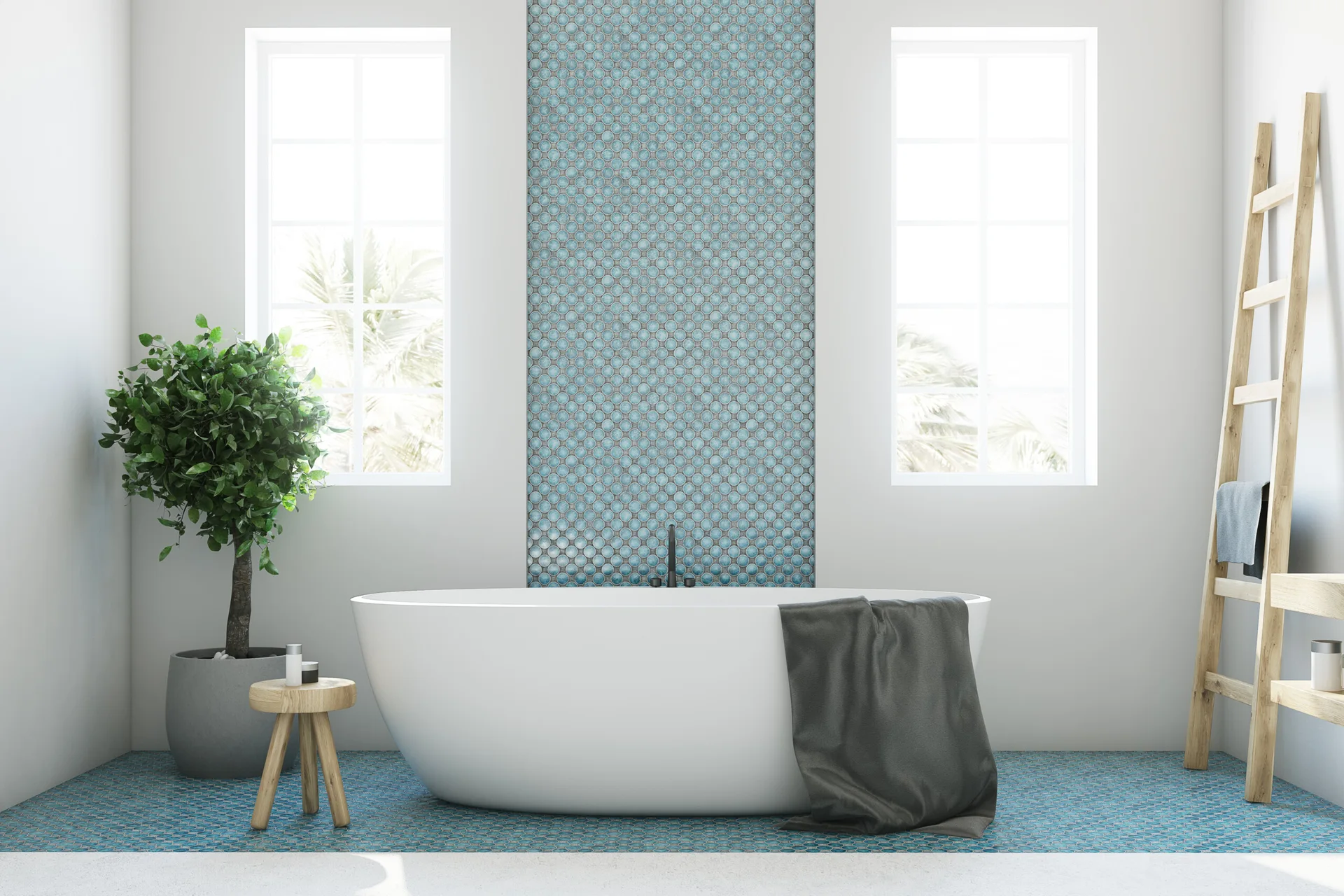 A white freestanding bathtub stands on blue mosaic floor tiles in front of a matching blue tile wall accent flanked by two tall windows.