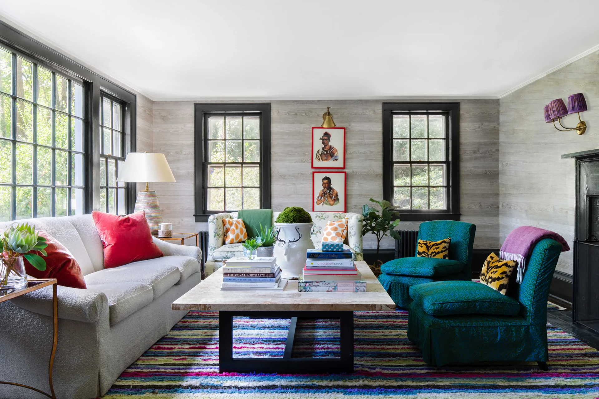 An eclectic living room features a cream sofa, patterned teal armchairs, a colorful striped rug, and black-trimmed windows against wood-grain wallpaper.