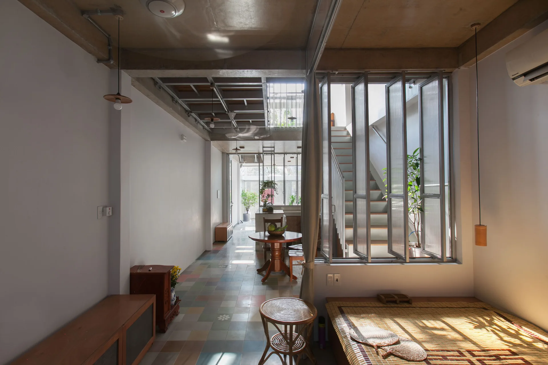An open residential interior featuring multicolored floor tiles, an exposed concrete ceiling, and a staircase behind a glass partition window.