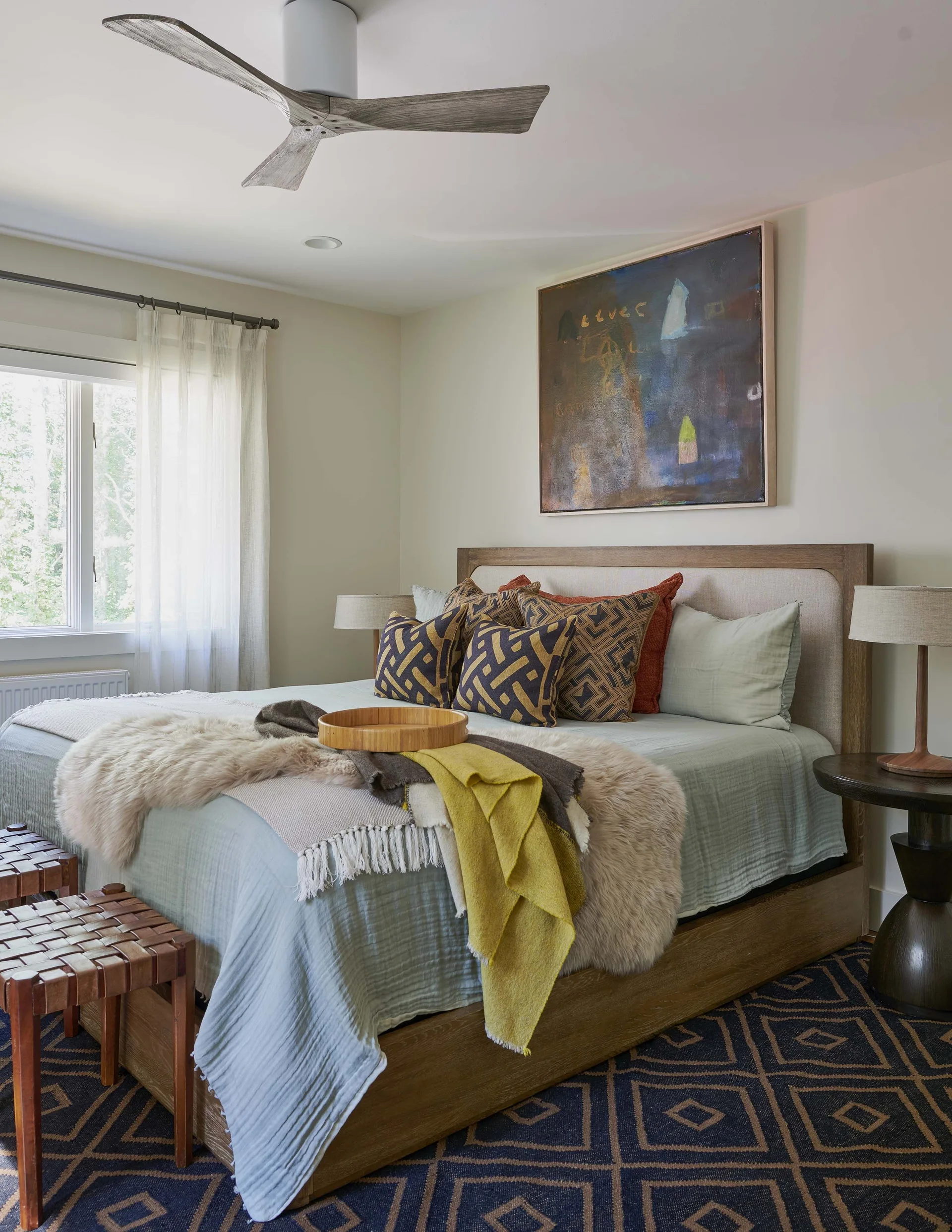 A bedroom featuring an upholstered wood bed frame with layered bedding, a navy geometric rug, and a large abstract painting.