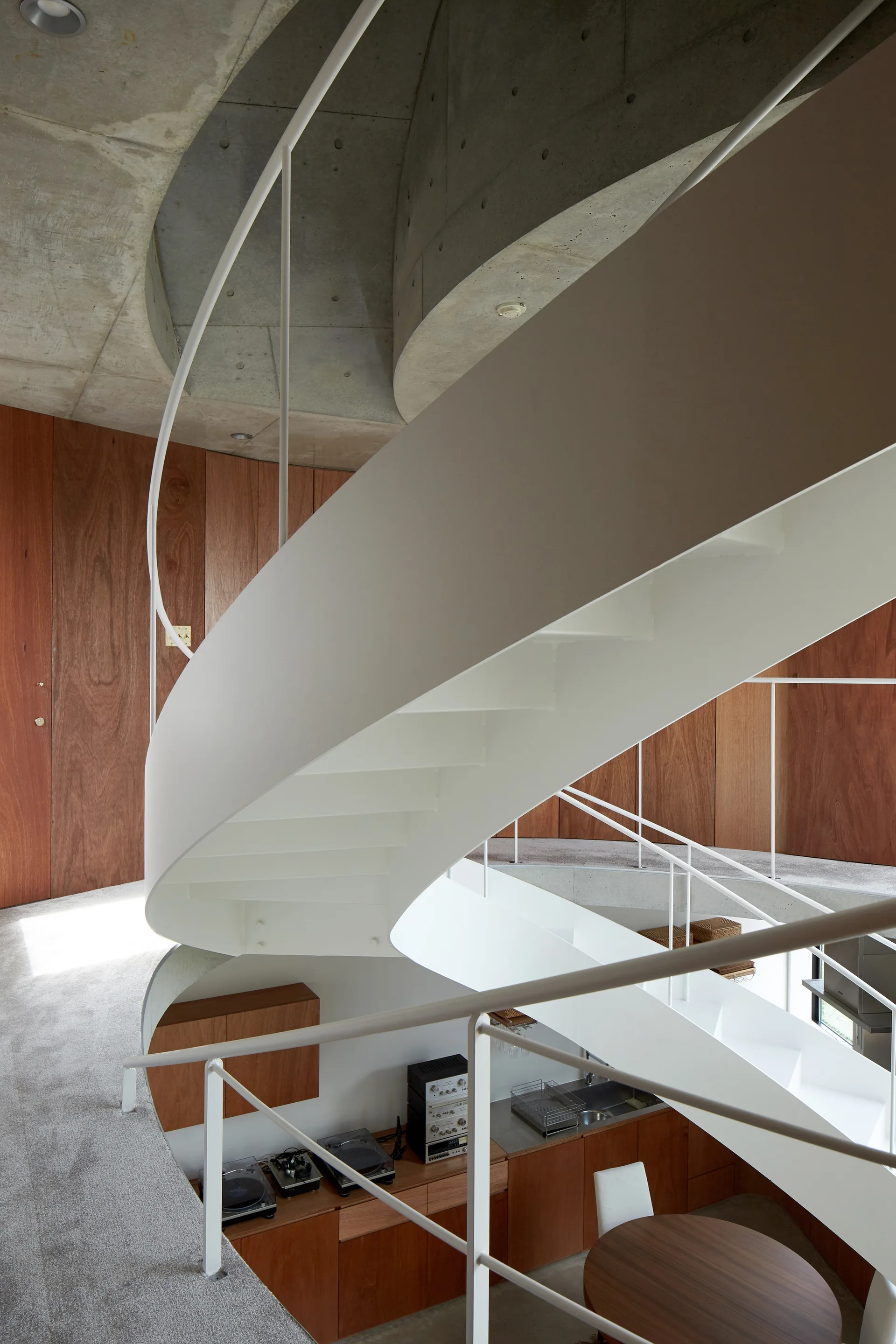 A curved white metal spiral staircase connects multiple levels in a modern room with warm wood-paneled walls and a raw concrete ceiling.