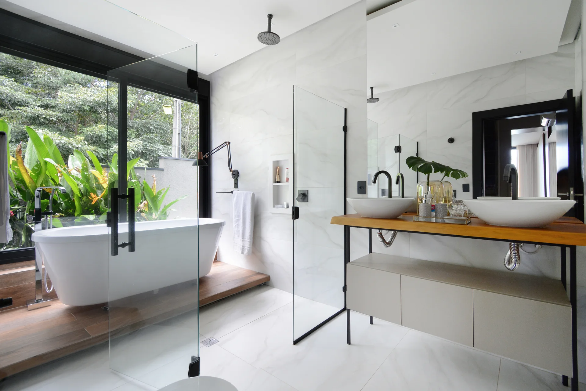 A modern bathroom features a freestanding bathtub on a wood platform next to a large window, a glass shower, and a wood double vanity.