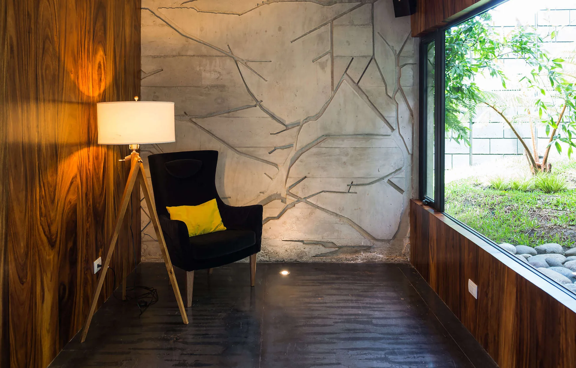 A dark armchair with a yellow pillow sits in a corner next to a wooden tripod lamp, flanked by a branch-textured concrete wall and wood paneling.