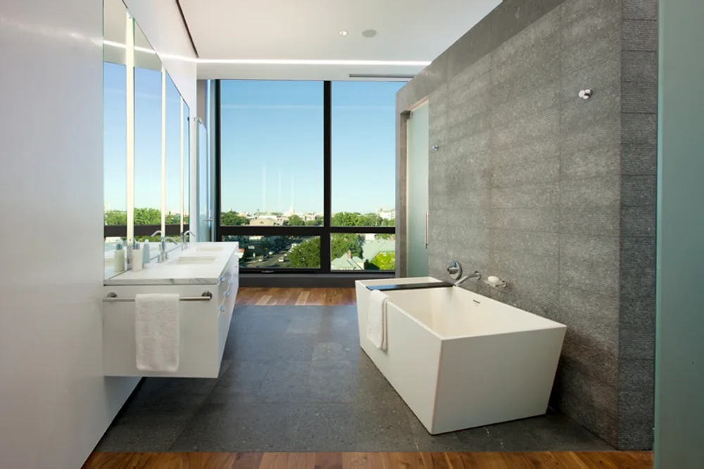 A contemporary bathroom features a white freestanding bathtub by a gray stone wall, a floating double vanity, and a large window with city views.