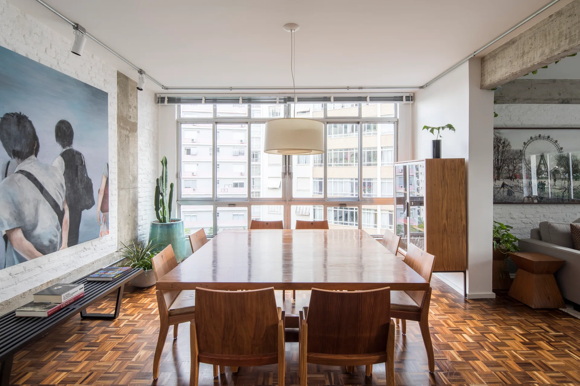 A spacious dining area features a square wooden table with matching chairs, polished parquet flooring, and a white painted brick wall.