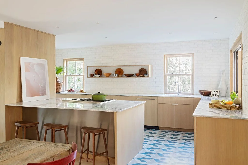 A modern kitchen features light wood cabinetry, stone countertops, wooden bar stools, white brick walls, and a blue and white geometric tile floor.