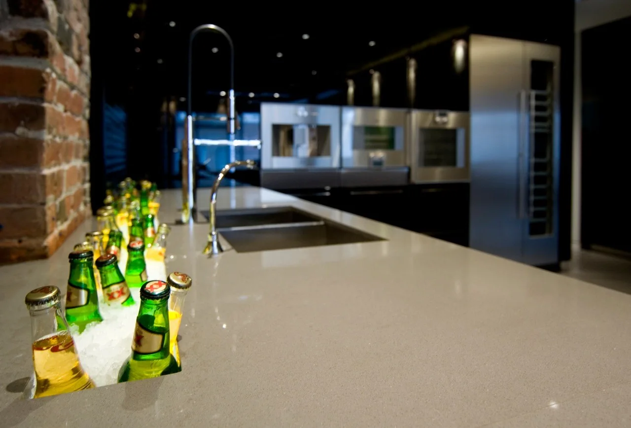 A modern dark kitchen island features an integrated narrow ice trough filled with green beverage bottles next to a stainless steel sink.