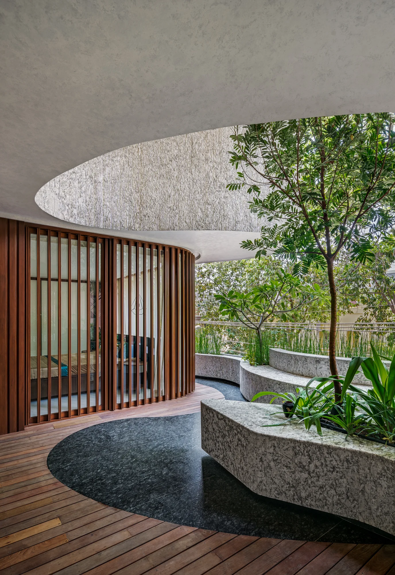 A modern outdoor terrace features curved wood-slatted doors, circular dark stone flooring inserts, wood decking, and built-in stone planters with trees.