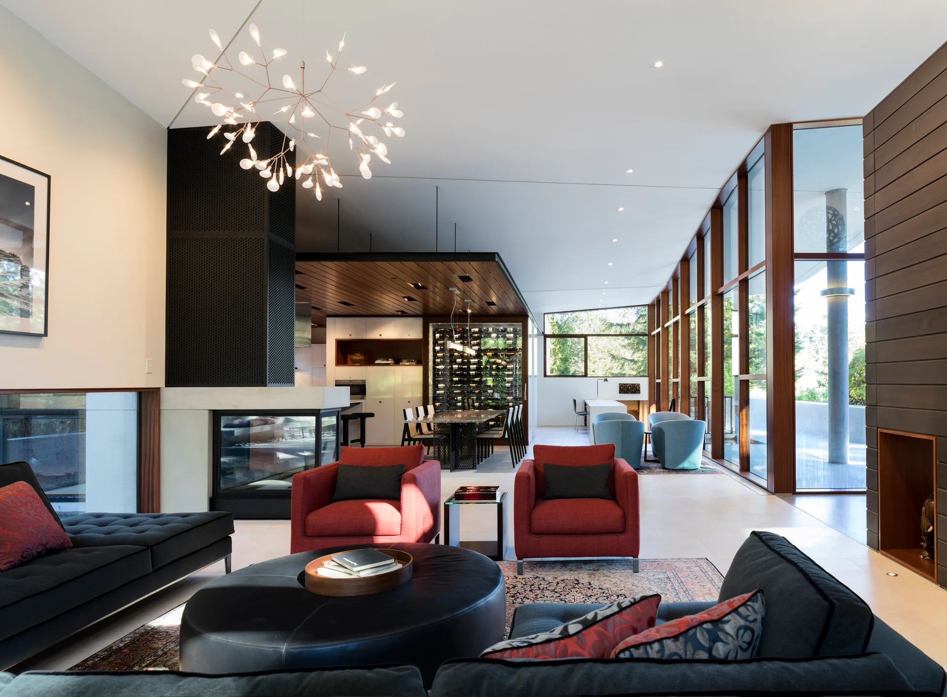 A modern open-concept living room features a pair of red armchairs, charcoal sofas, and a tall black fireplace next to wood-framed windows.