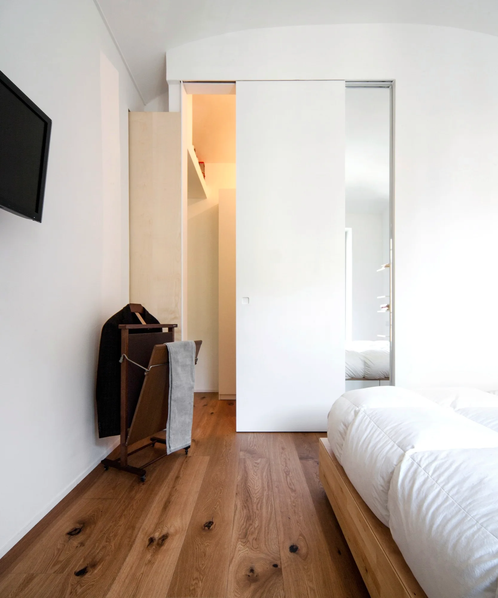 A minimalist bedroom features a low wood platform bed, wide-plank wood flooring, and a large white sliding closet door next to a vertical mirror.