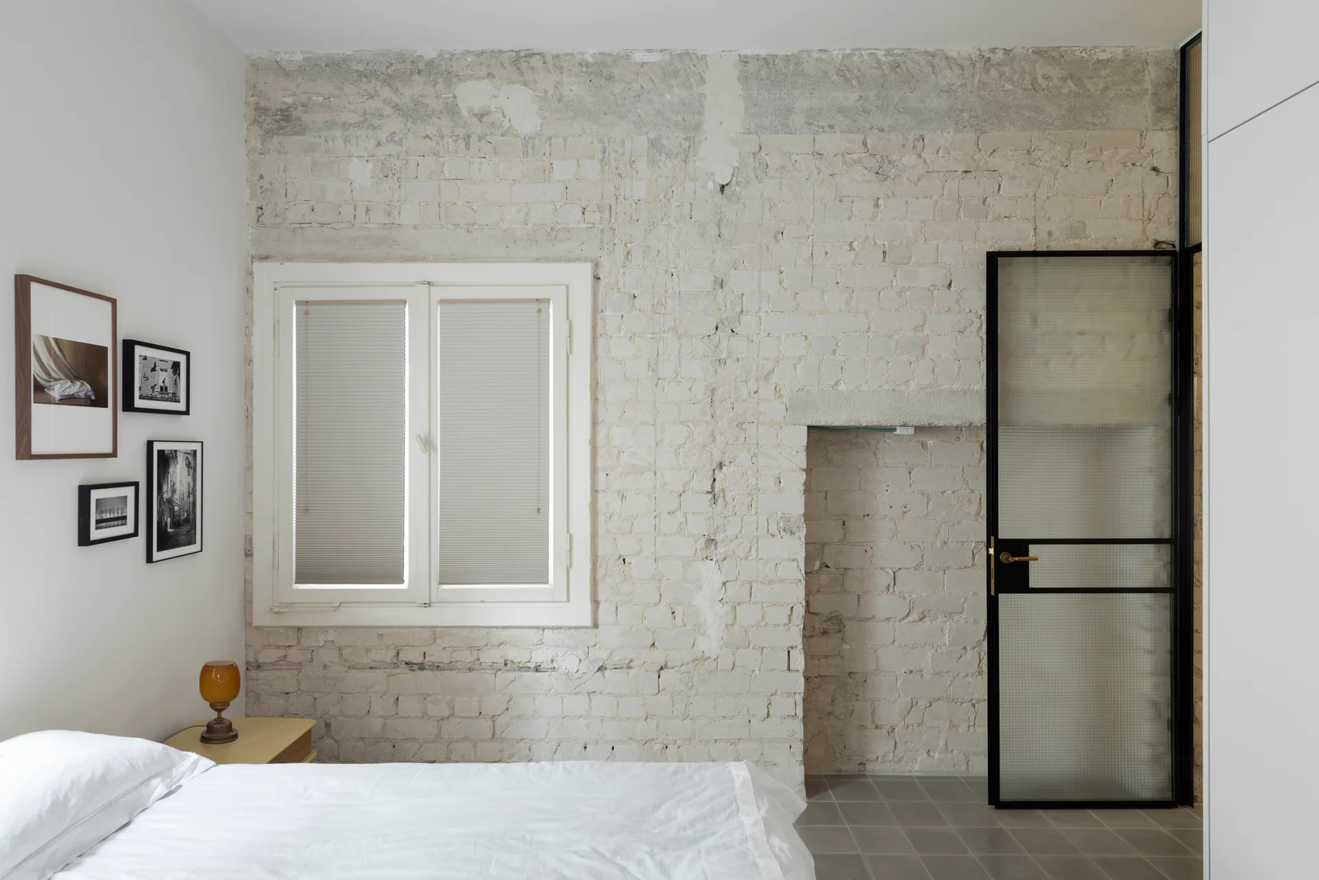 A minimalist bedroom featuring an exposed off-white brick wall, a window with pleated blinds, and a black-framed textured glass door.