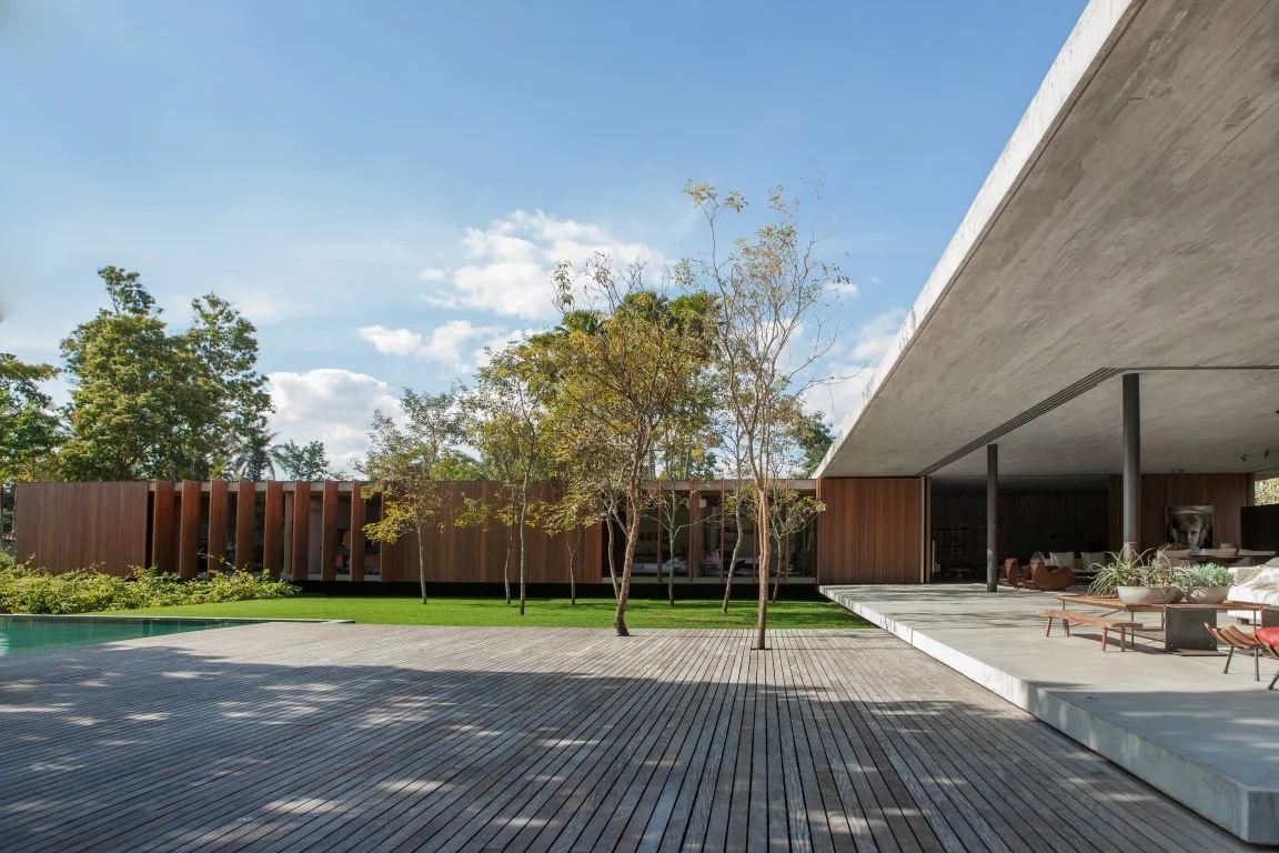 A modern outdoor pavilion features an expansive cantilevered concrete roof, a weathered wood deck, a lawn, and vertical wood-slat walls next to a pool.