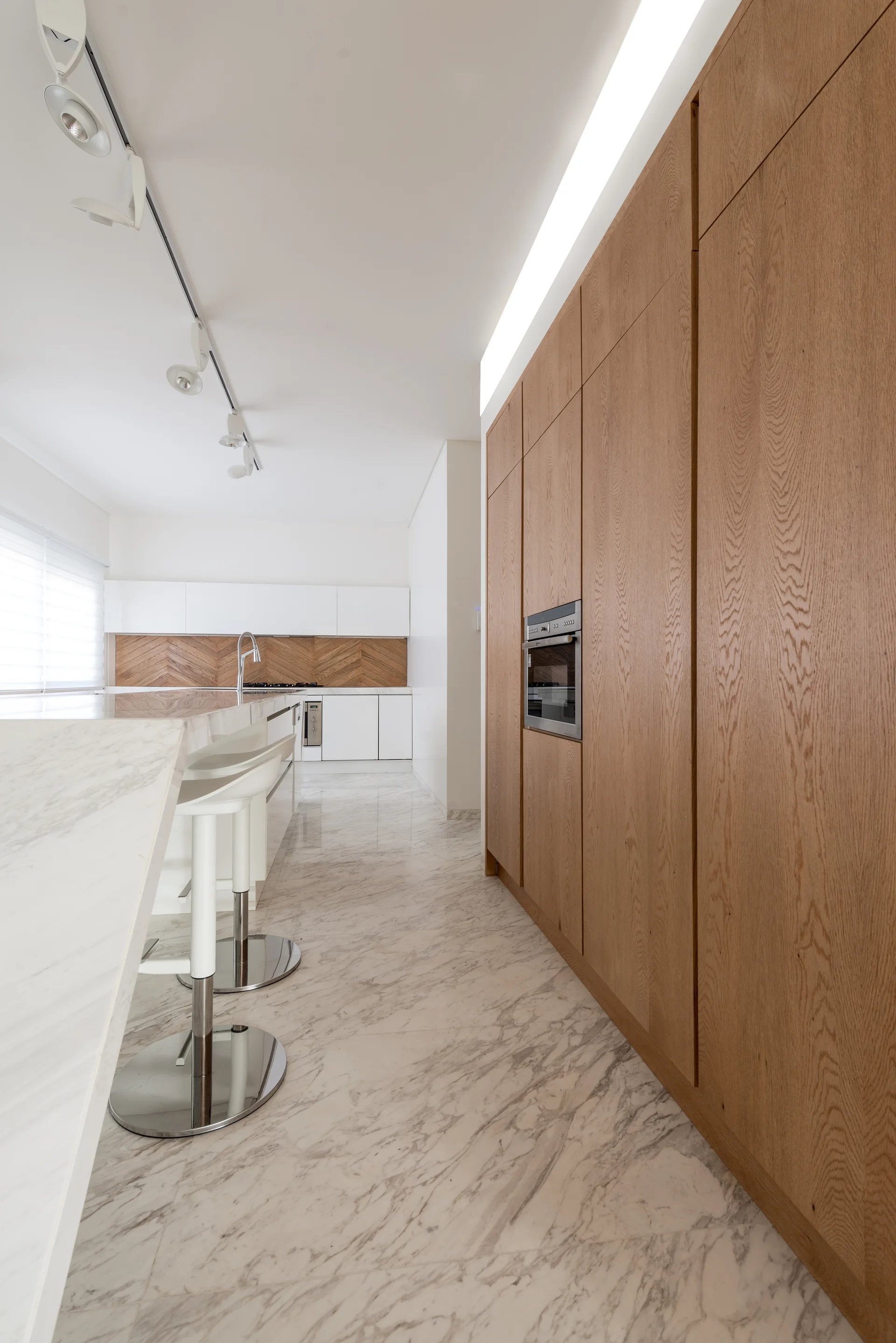 A modern kitchen featuring a towering wall of oak cabinets, polished white marble floor tiles, and a marble island with chrome bar stools.