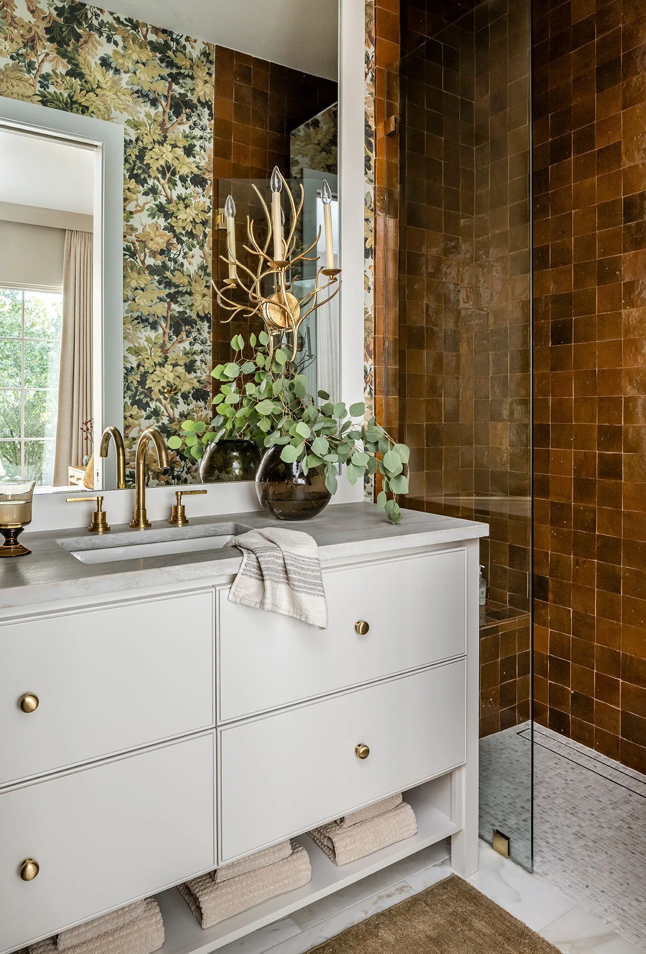 A white bathroom vanity with brass fixtures stands next to a walk-in shower with glossy brown tiles, backed by green leafy wallpaper.