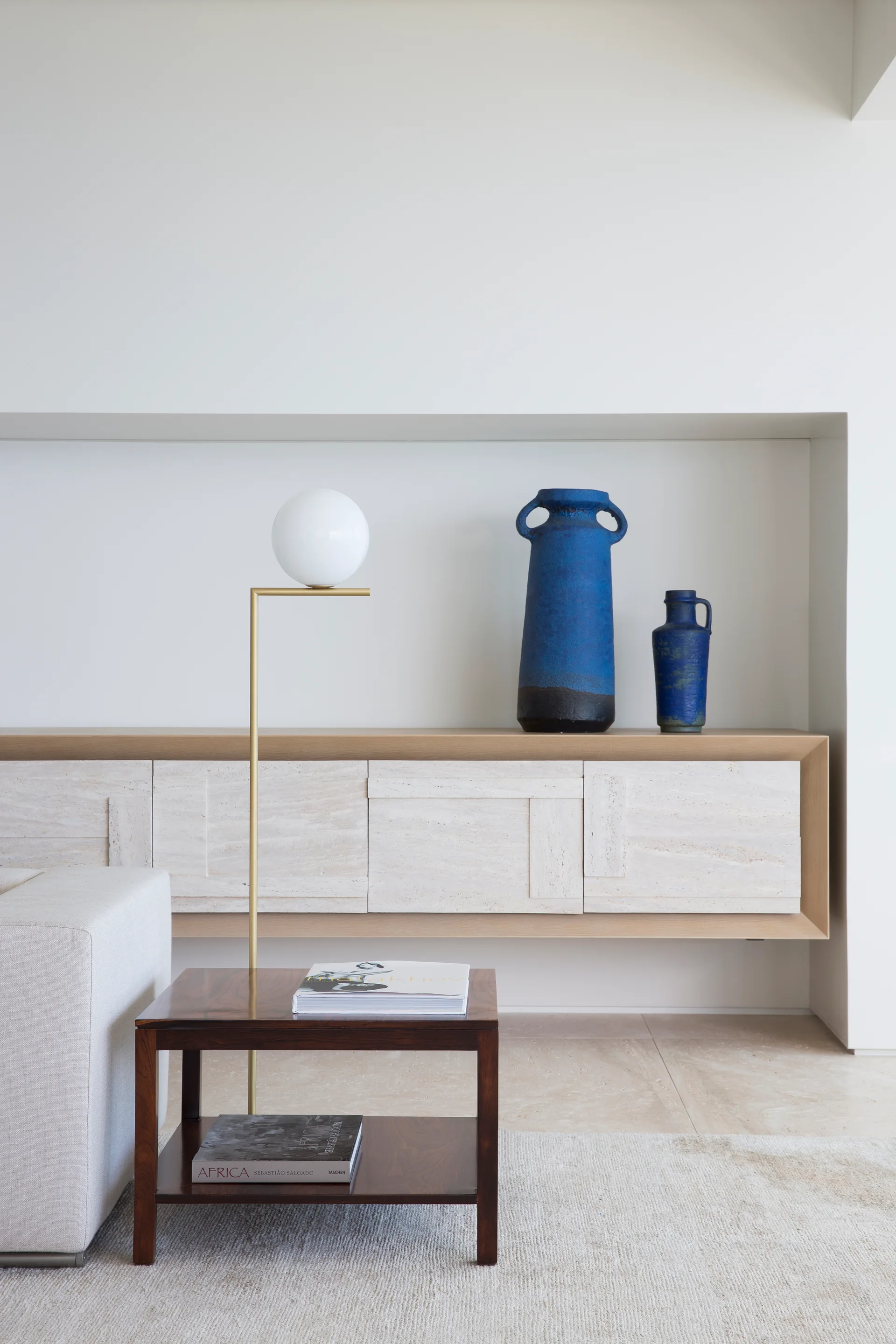 A floating travertine console inside a wall recess holds two cobalt blue vases next to a brass floor lamp and a dark wood side table.