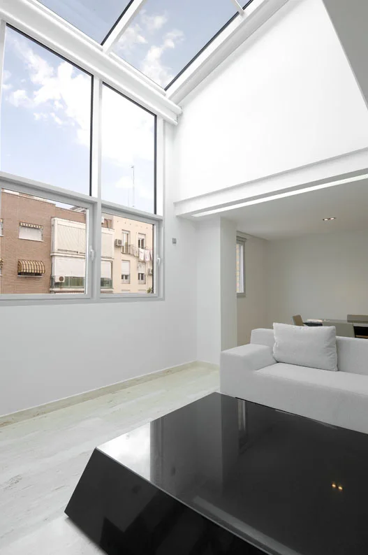 A bright minimalist living room featuring a double-height ceiling, large skylight windows, a light grey sofa, and a black glossy coffee table.