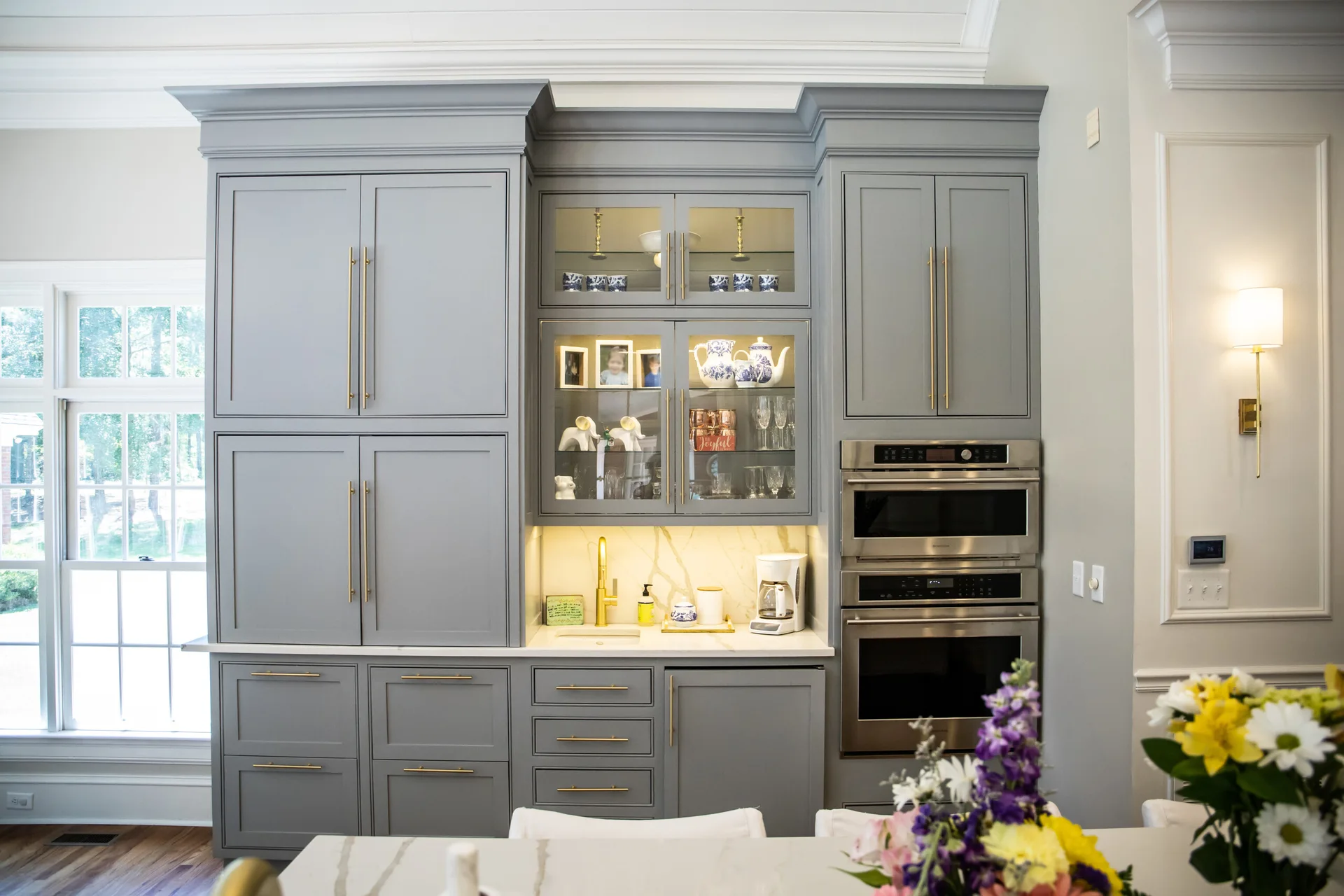 A large wall of grey kitchen cabinetry featuring a built-in double oven, glass-front display cases, brass hardware, and a small countertop wet bar.