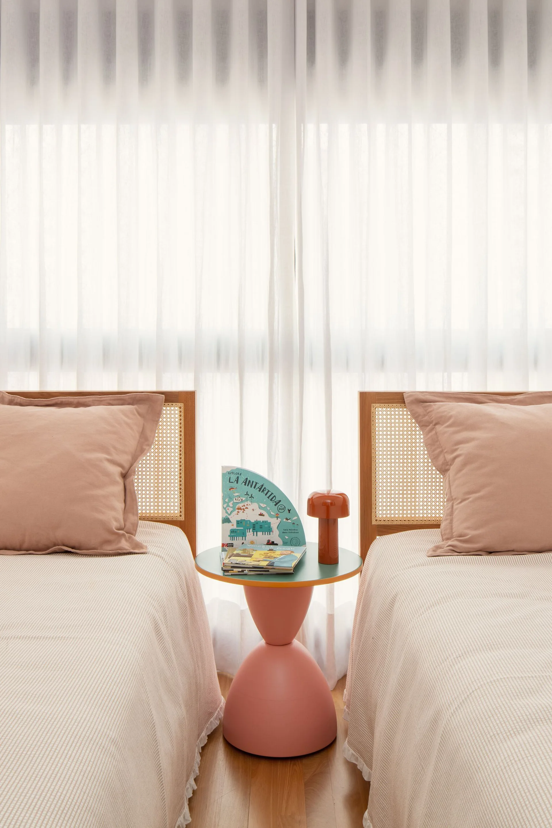 Two twin beds with woven cane headboards and pink pillows sit symmetrically on either side of a pink sculptural side table against sheer white curtains.