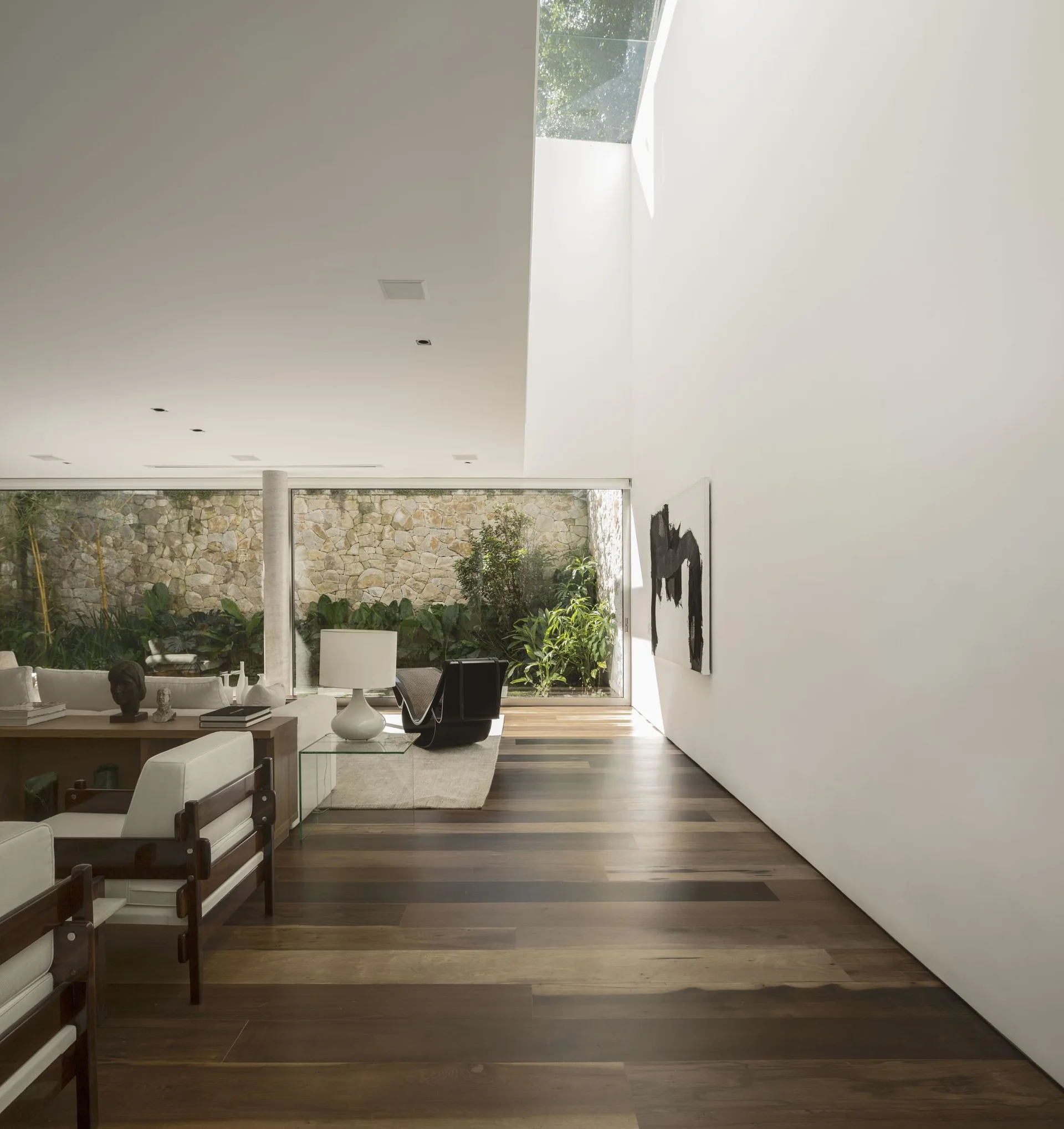 A minimalist living room with white walls, dark wood floors, a skylight, and large windows looking out to a stone-walled courtyard.