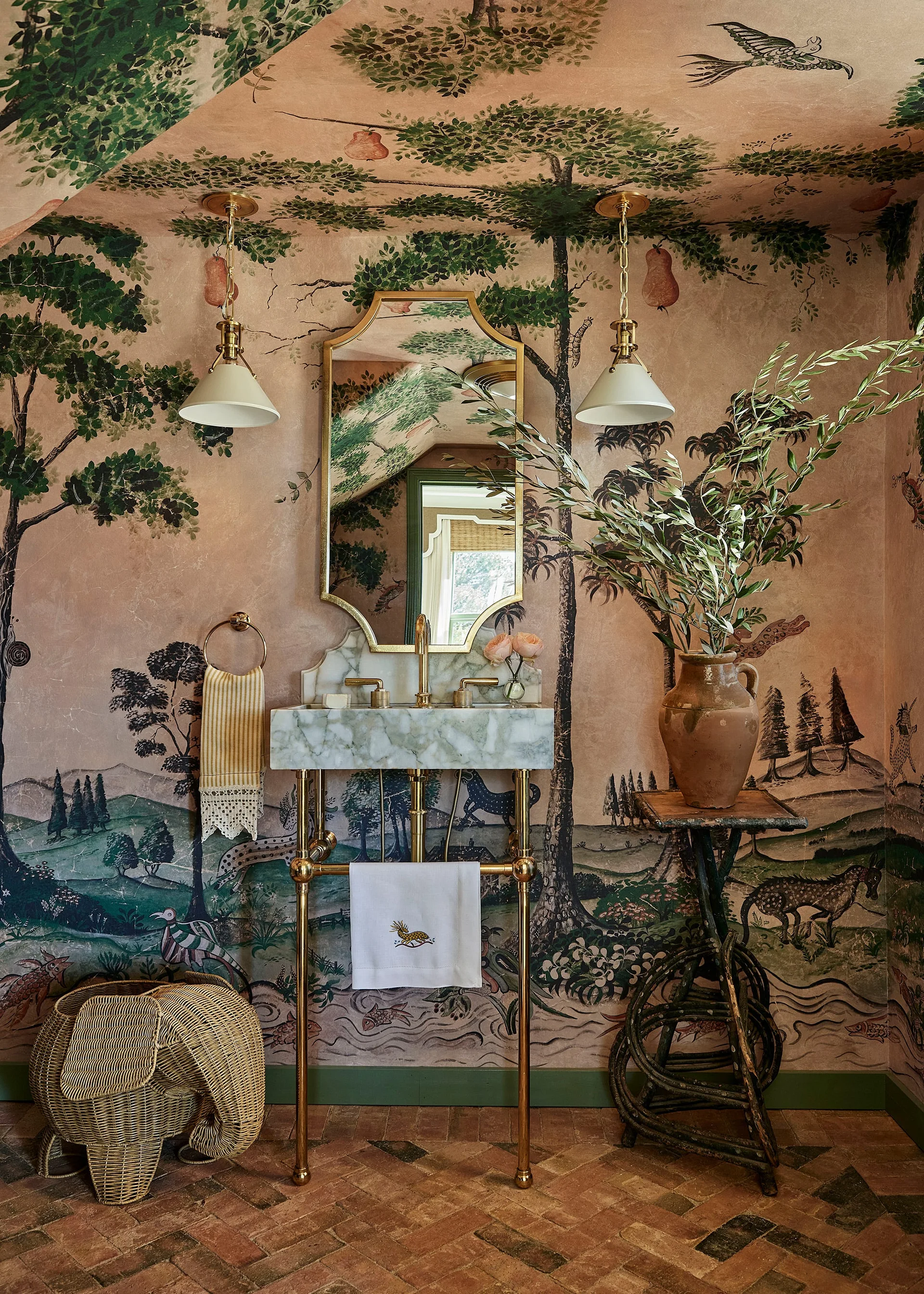 A powder room features scenic pink and green mural wallpaper, a marble washstand with brass legs, and terracotta brick tile floors.