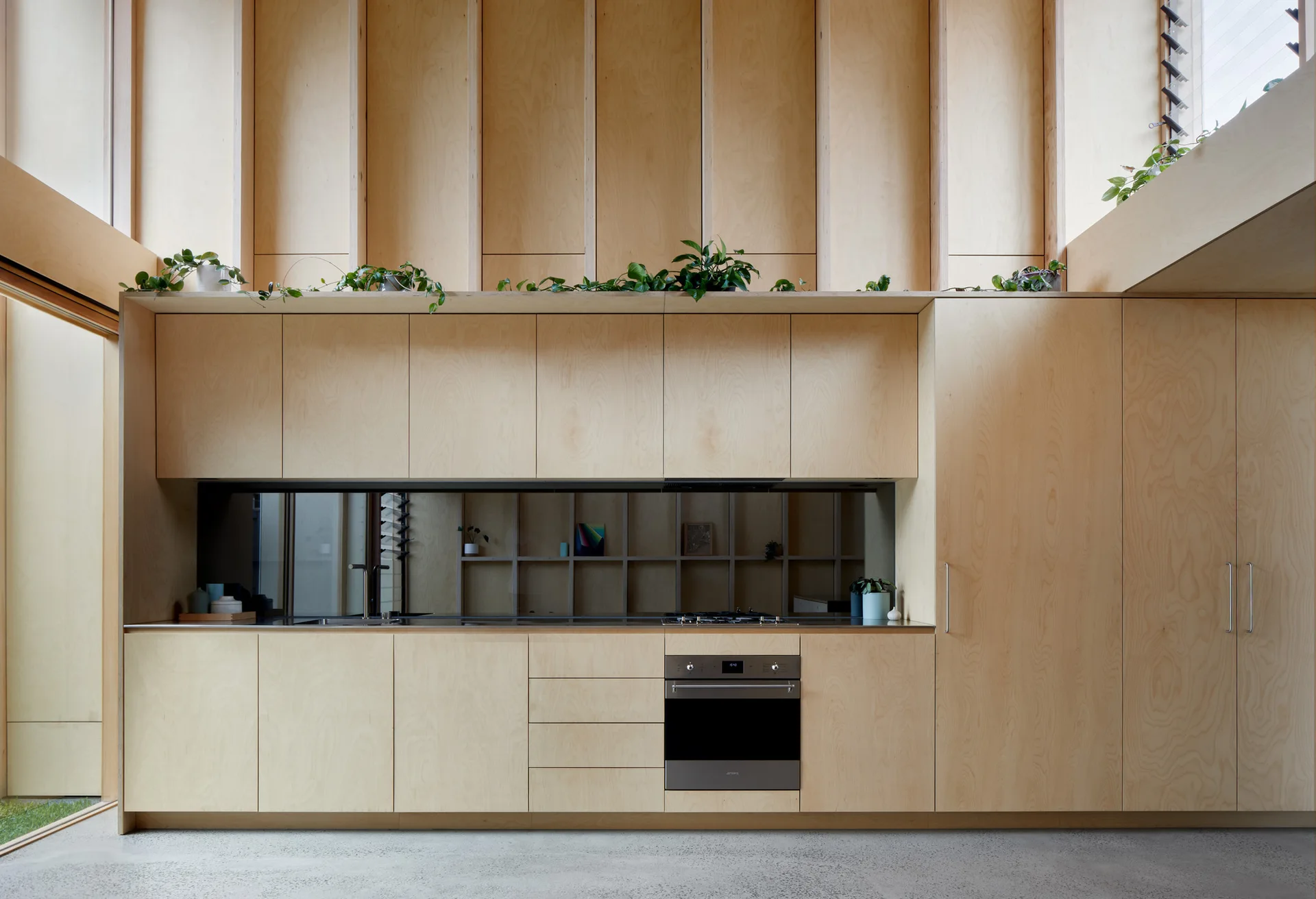 A minimalist kitchen features seamless light wood plywood cabinets, an integrated stainless steel oven, and a dark reflective glass backsplash.