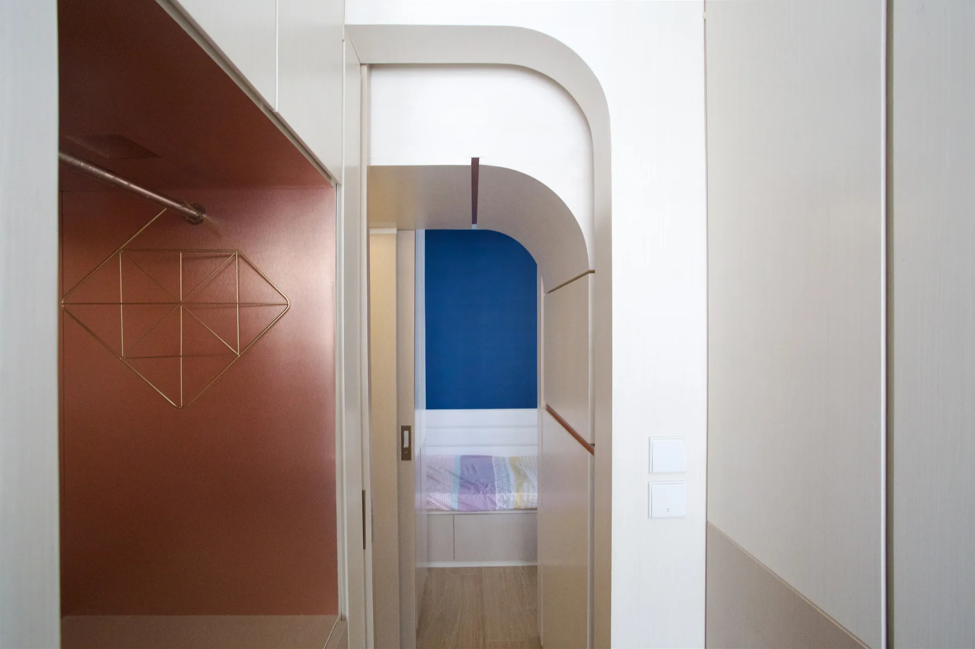 A view through a rounded white archway showing a bedroom with a blue wall, adjacent to an integrated closet niche with copper metallic walls.