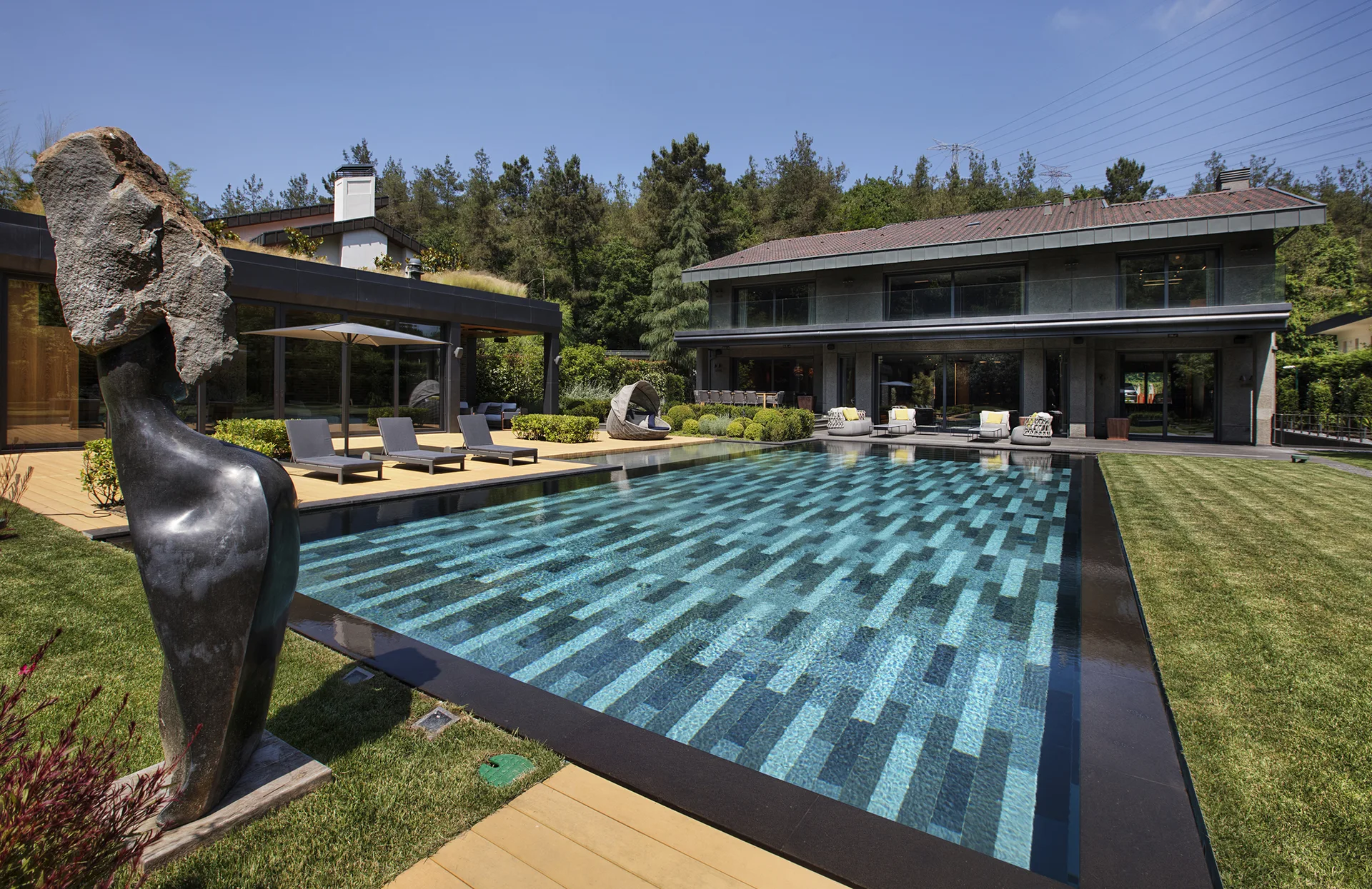 A large backyard swimming pool with patterned blue and gray tiling, adjacent to a wooden deck, manicured lawn, and modern two-story home.