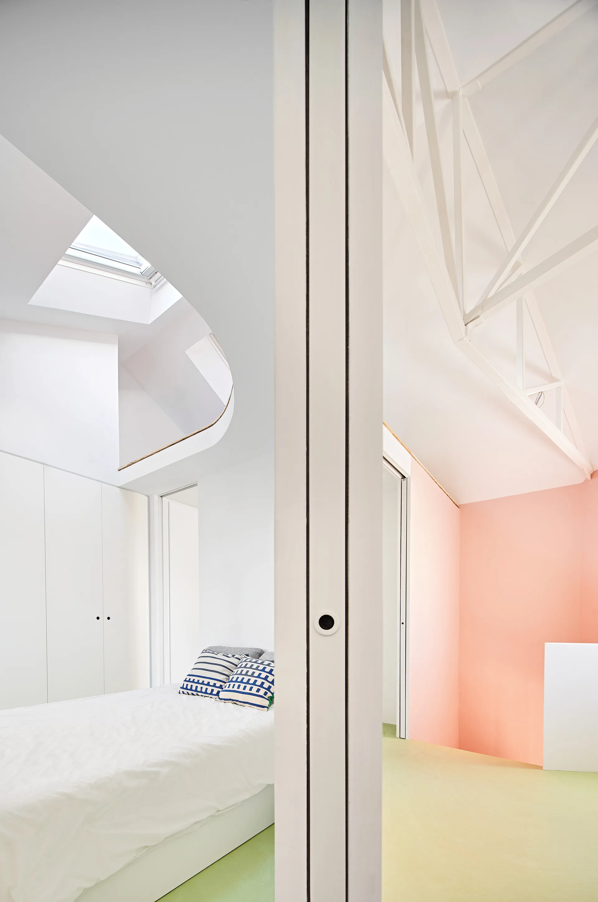 A modern bedroom features a white bed with striped pillows, a skylight, a sliding door partition, a pastel pink wall, and white ceiling beams.