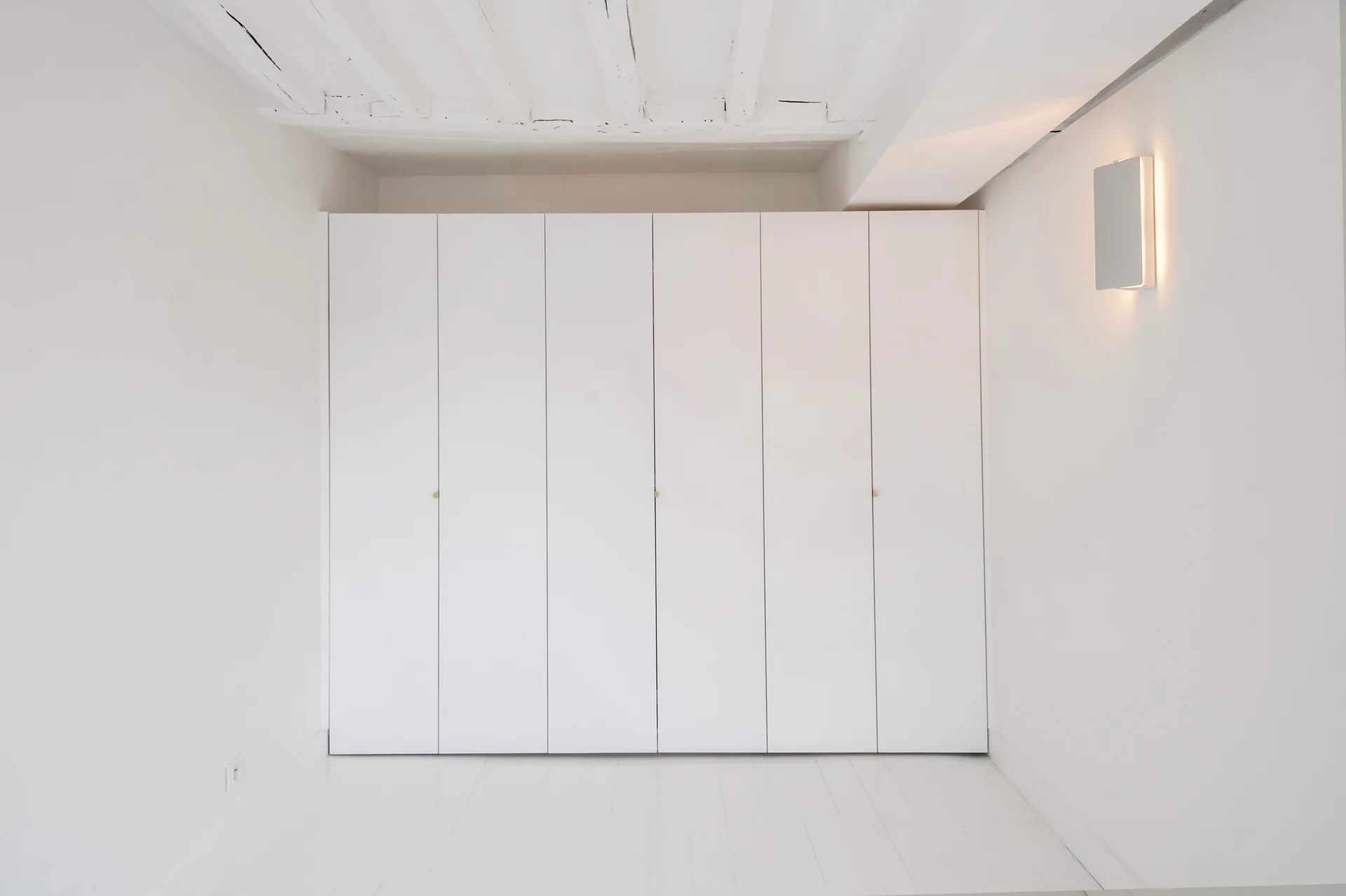 Floor-to-ceiling white wardrobe cabinet built beneath painted wooden ceiling beams in a minimalist room with a modern rectangular wall sconce.