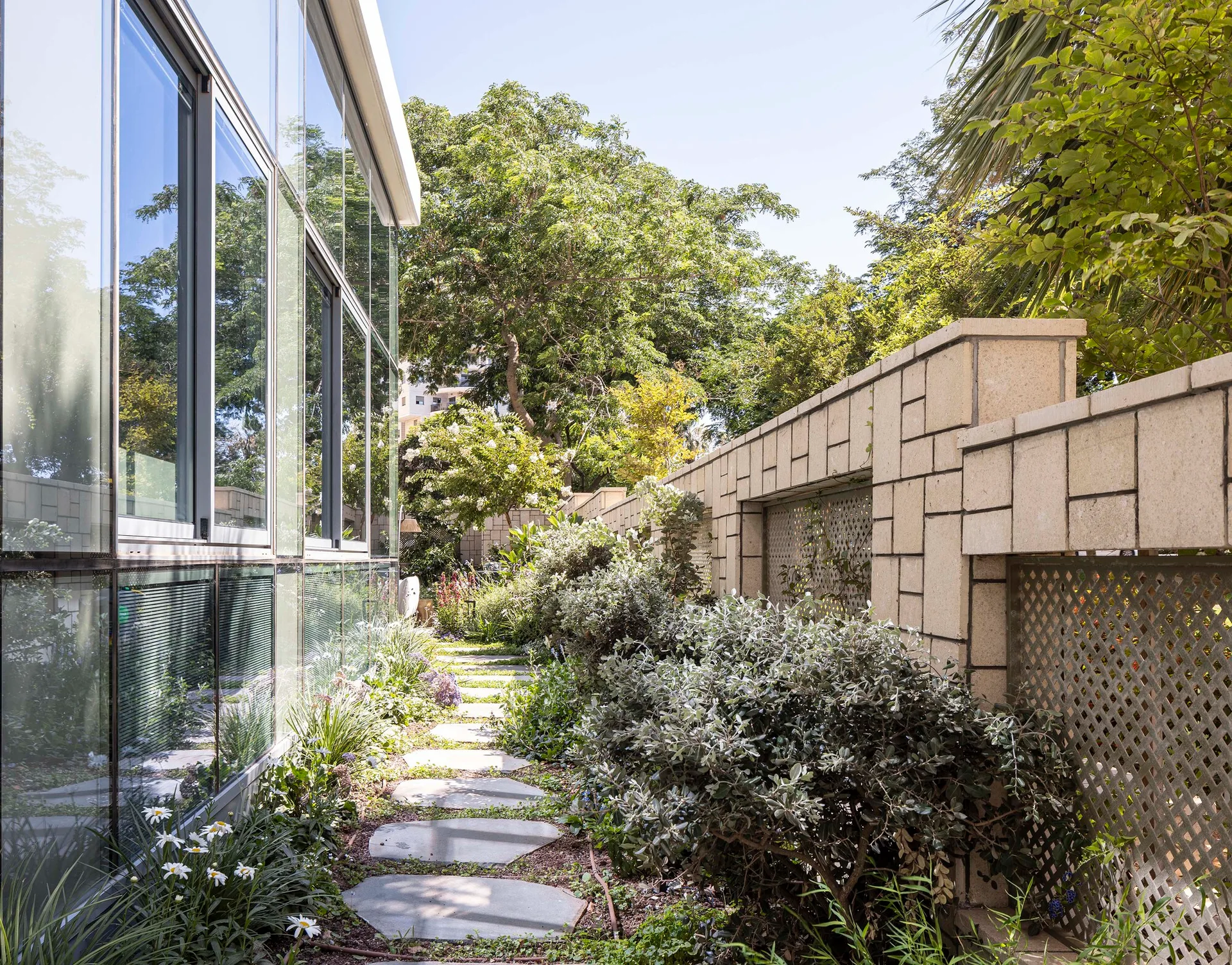 A narrow outdoor garden path with irregular stone stepping stones bordered by a reflective glass building facade and a tiered stone wall with lattice panels.
