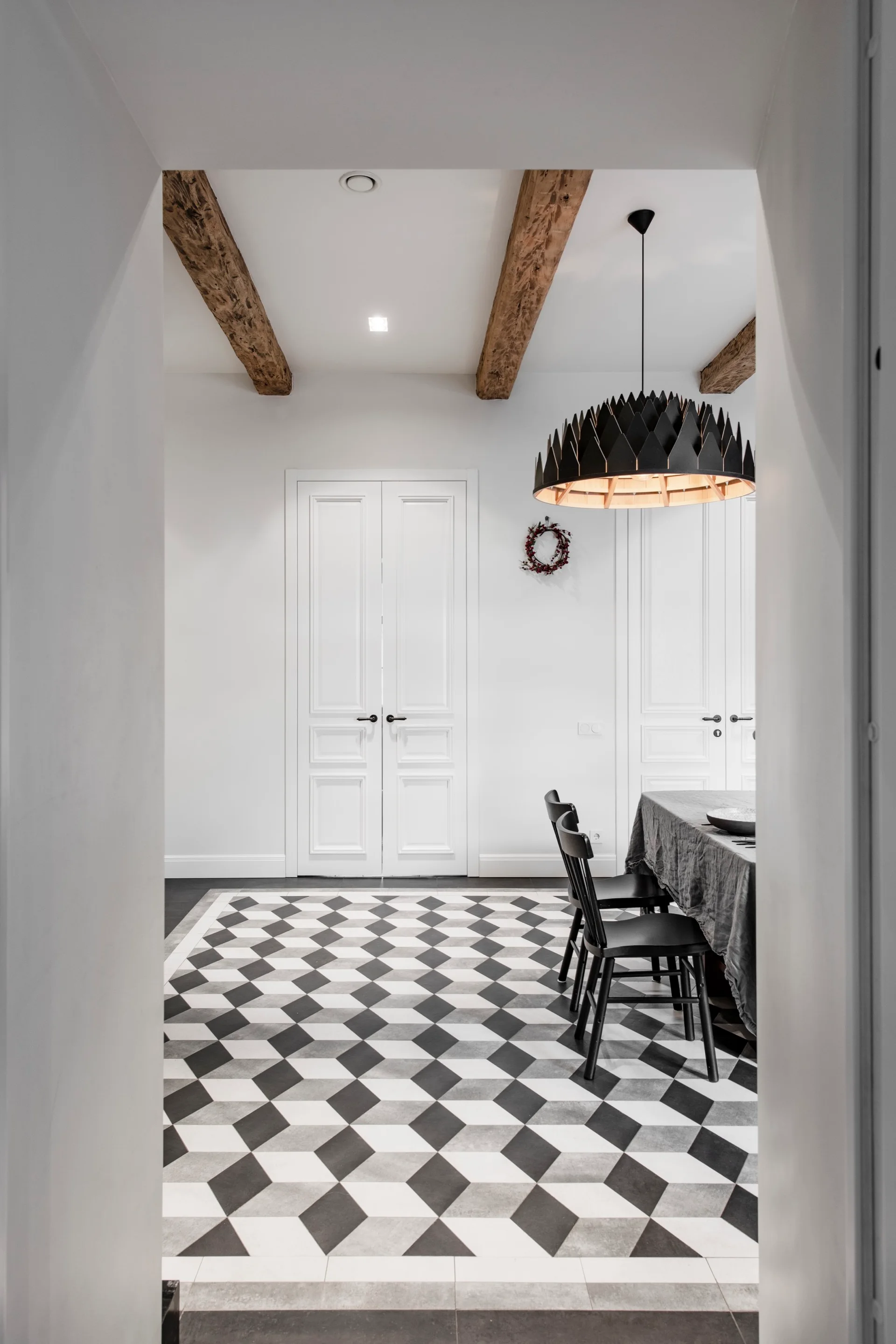 A dining room viewed through an archway featuring black and white geometric floor tiles, a dining table, black chairs, and exposed wood ceiling beams.