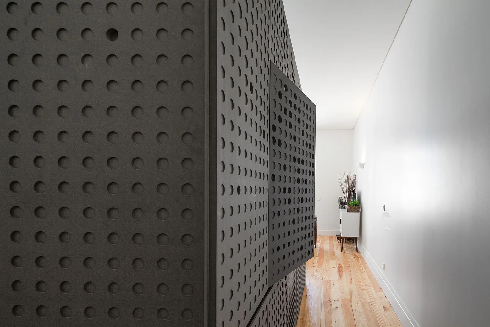 A large charcoal grey perforated partition with a pattern of circular cutouts stands in a minimalist hallway with light wood floors.