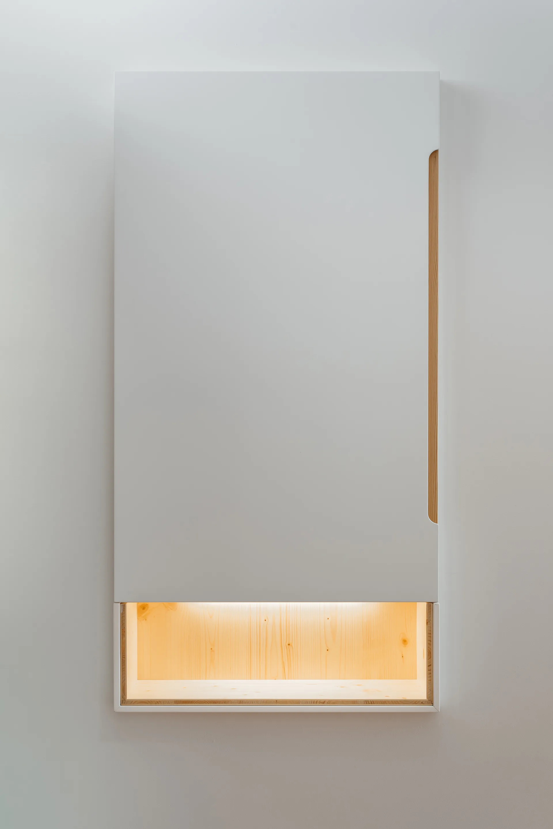 A vertical white wall cabinet featuring a minimalist recessed wood shelf with warm integrated lighting at the bottom and a slim wood handle.