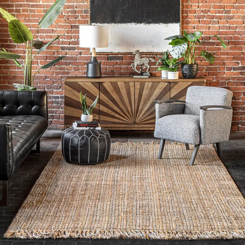 A living room with an exposed brick wall features a wooden sunburst console, a gray armchair, a black leather pouf, and a textured woven rug.