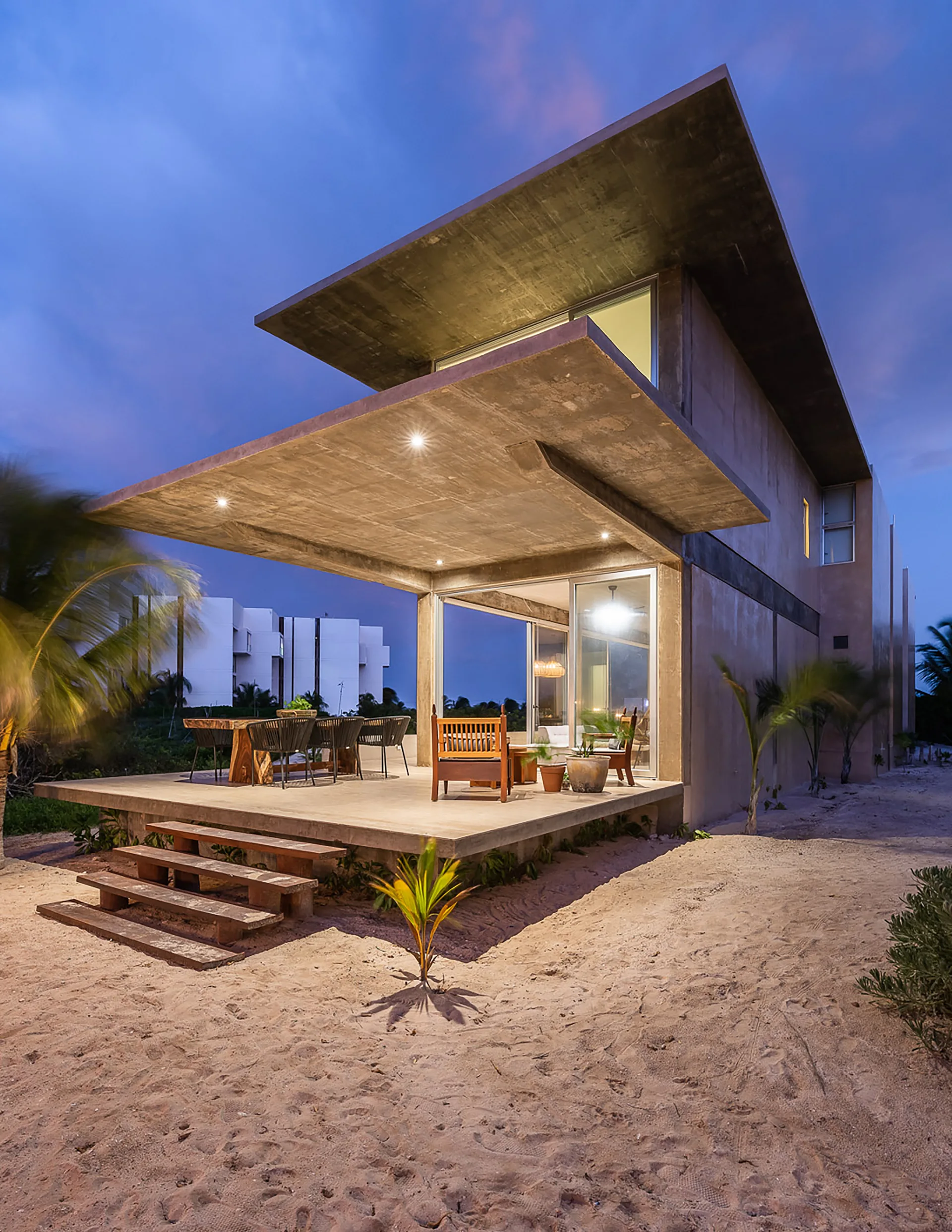 A modern concrete patio with a dramatic cantilevered roof overhang features an outdoor dining set, wooden chairs, and low steps leading to sandy ground.
