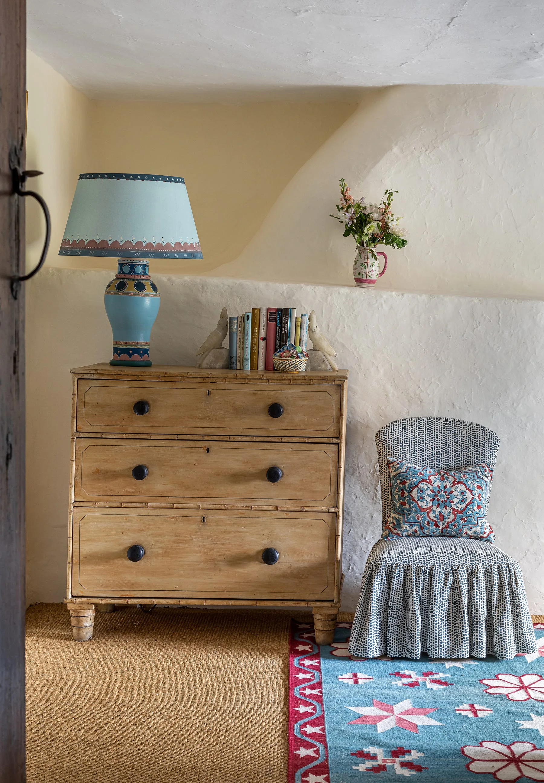 A light wood chest of drawers with a blue hand-painted lamp sits beside a patterned skirted chair against textured plaster walls.